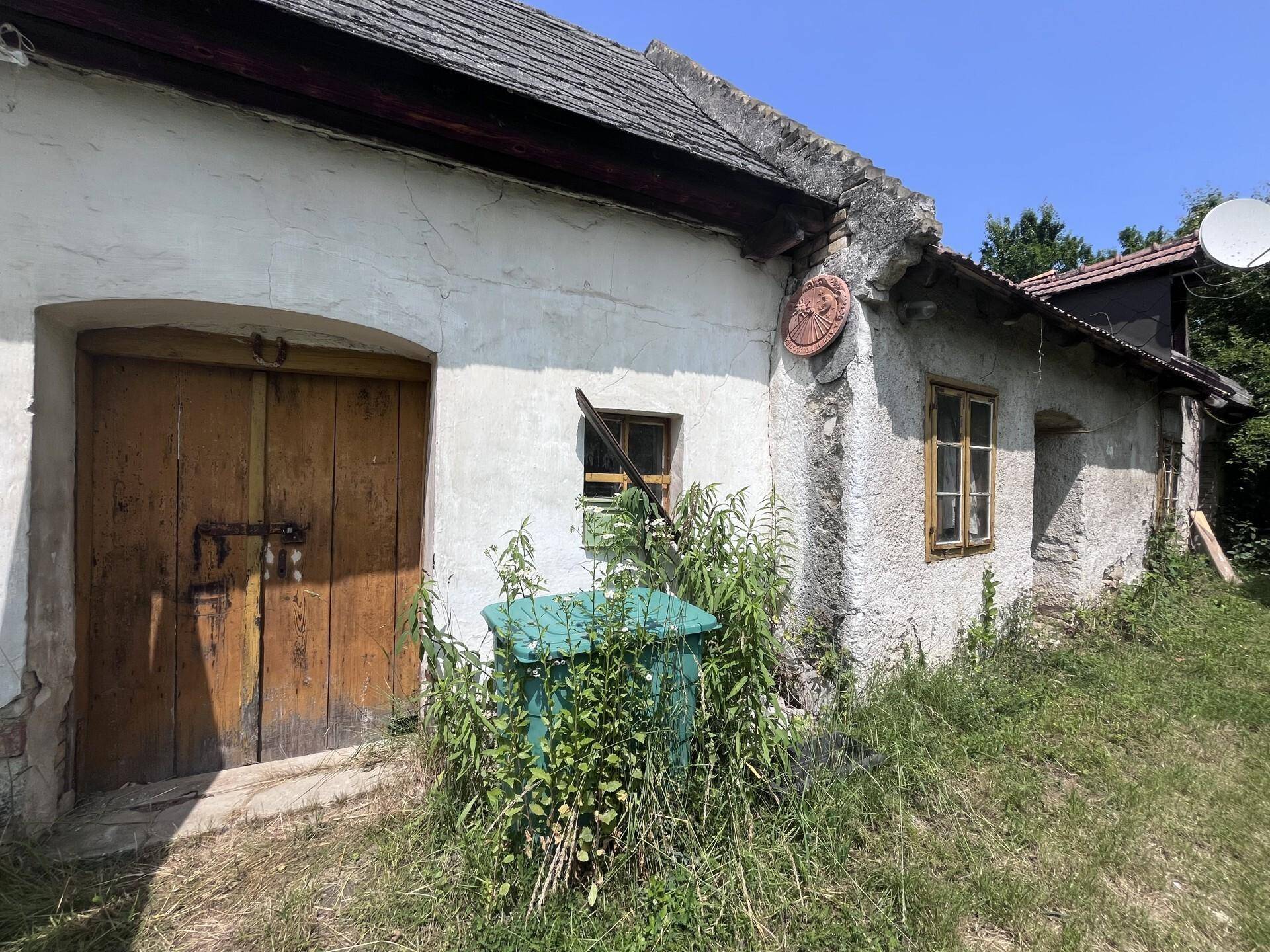 Rustikales altes Gebäude mit Holztür, kleinen Fenstern und überwuchertem Gelände.