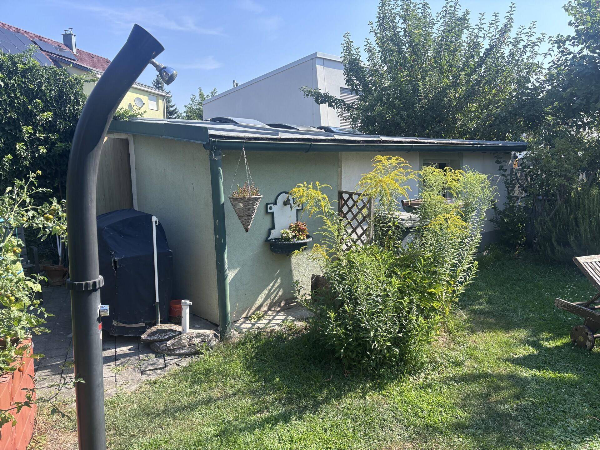 Gartenbereich mit einem Geräteschuppen, einer Außendusche und üppiger Vegetation unter blauem Himmel.
