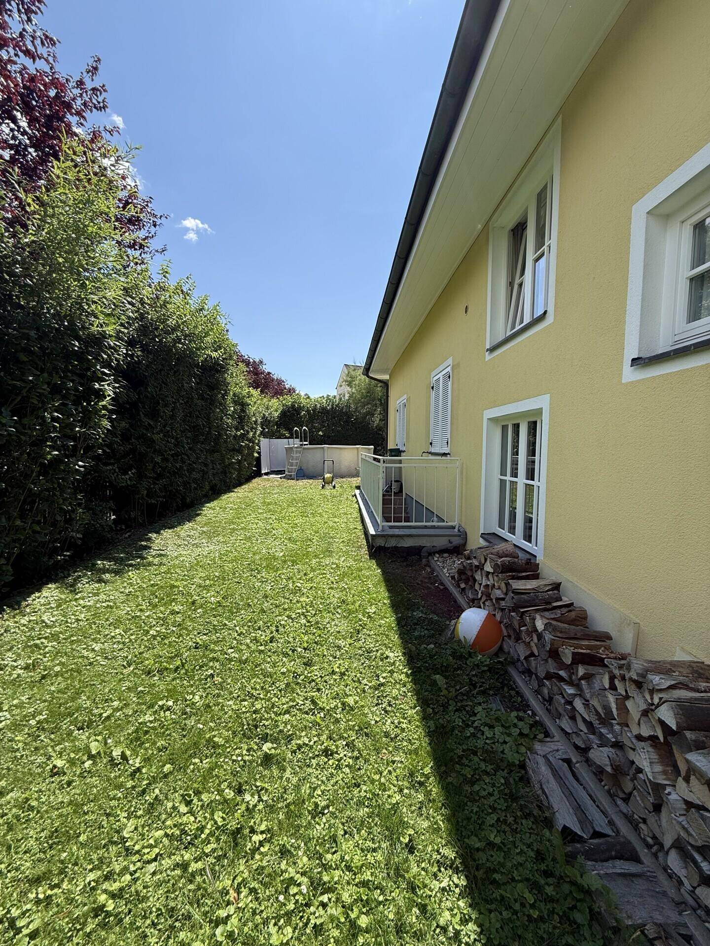 Seitlicher Gartenbereich mit Rasenfläche, Holzstapel und Blick auf den Pool im Hintergrund.