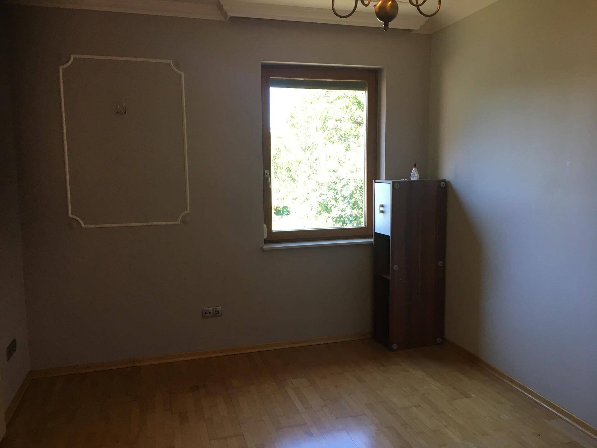 Leeres Zimmer mit hellgrauen Wänden, Fenster und Holzparkettboden.
