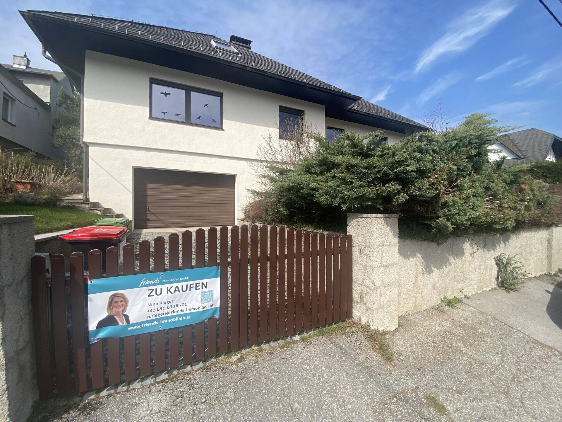 Vorderansicht des Hauses mit Garage, Einfahrt und einem Maklerschild am Holzzaun.