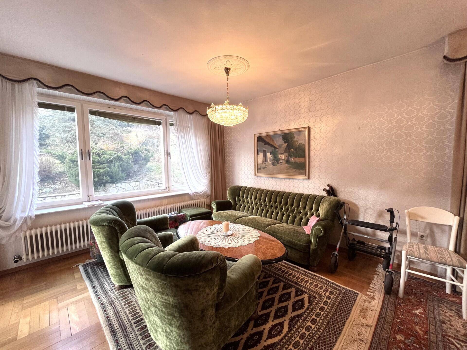 Wohnzimmer mit grünen Samtsofas, Couchtisch und großem Fenster mit Blick ins Grüne.