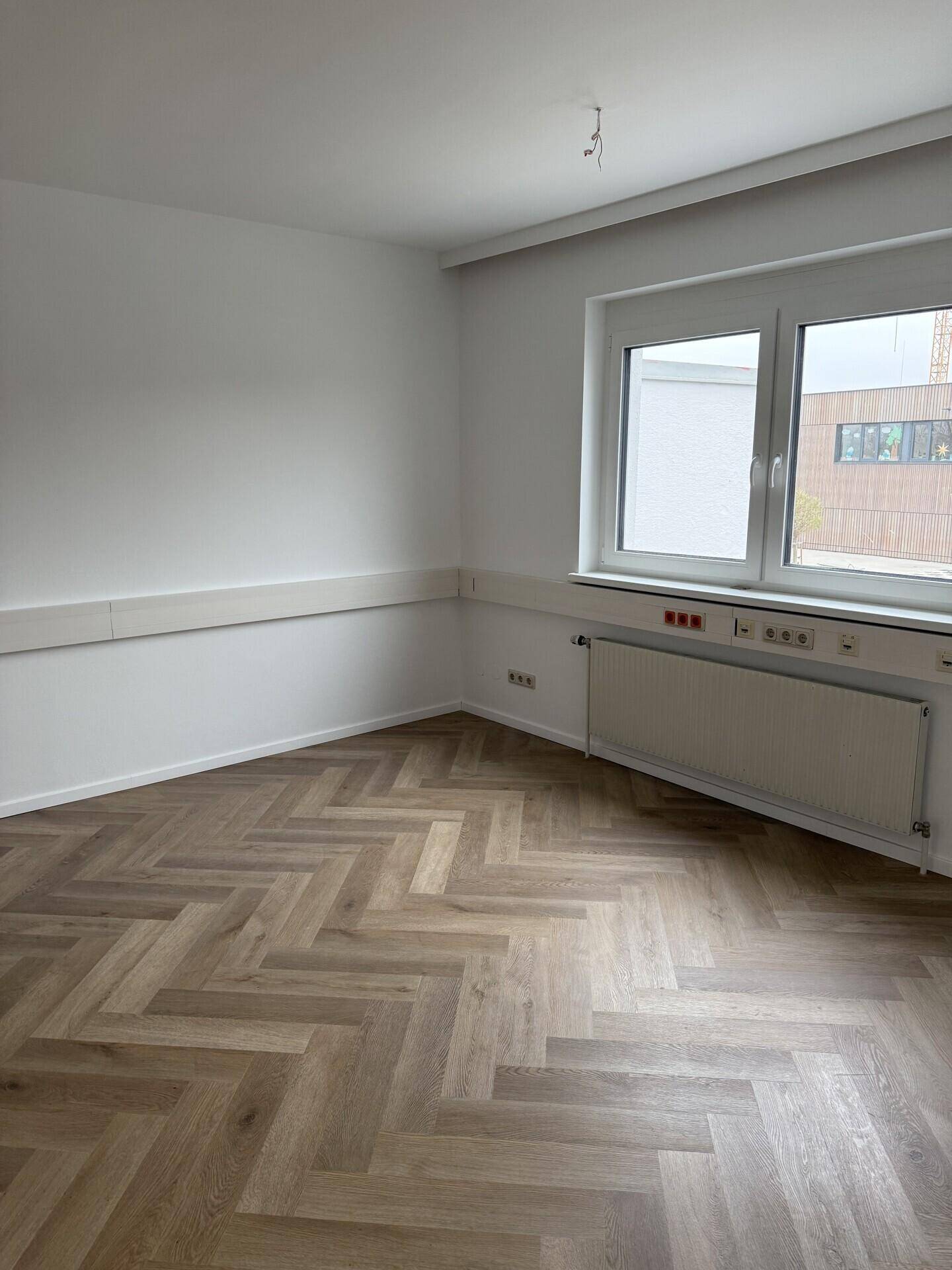 Heller, leerer Büroraum mit Fischgrätparkett, einem Fenster und einem Heizkörper an der Wand.
