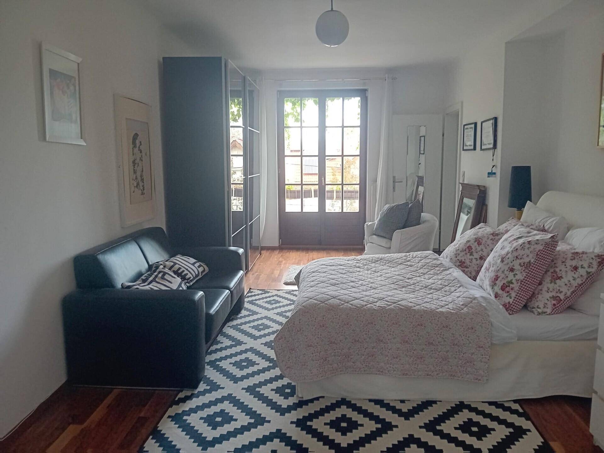 Helles Schlafzimmer mit Doppelbett, Sofa und großem Fenster, Holzboden.