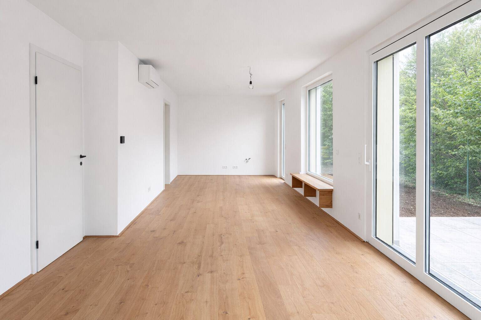 Helles, leeres Wohnzimmer mit Holzboden, großen Fenstern und Zugang zum Garten.