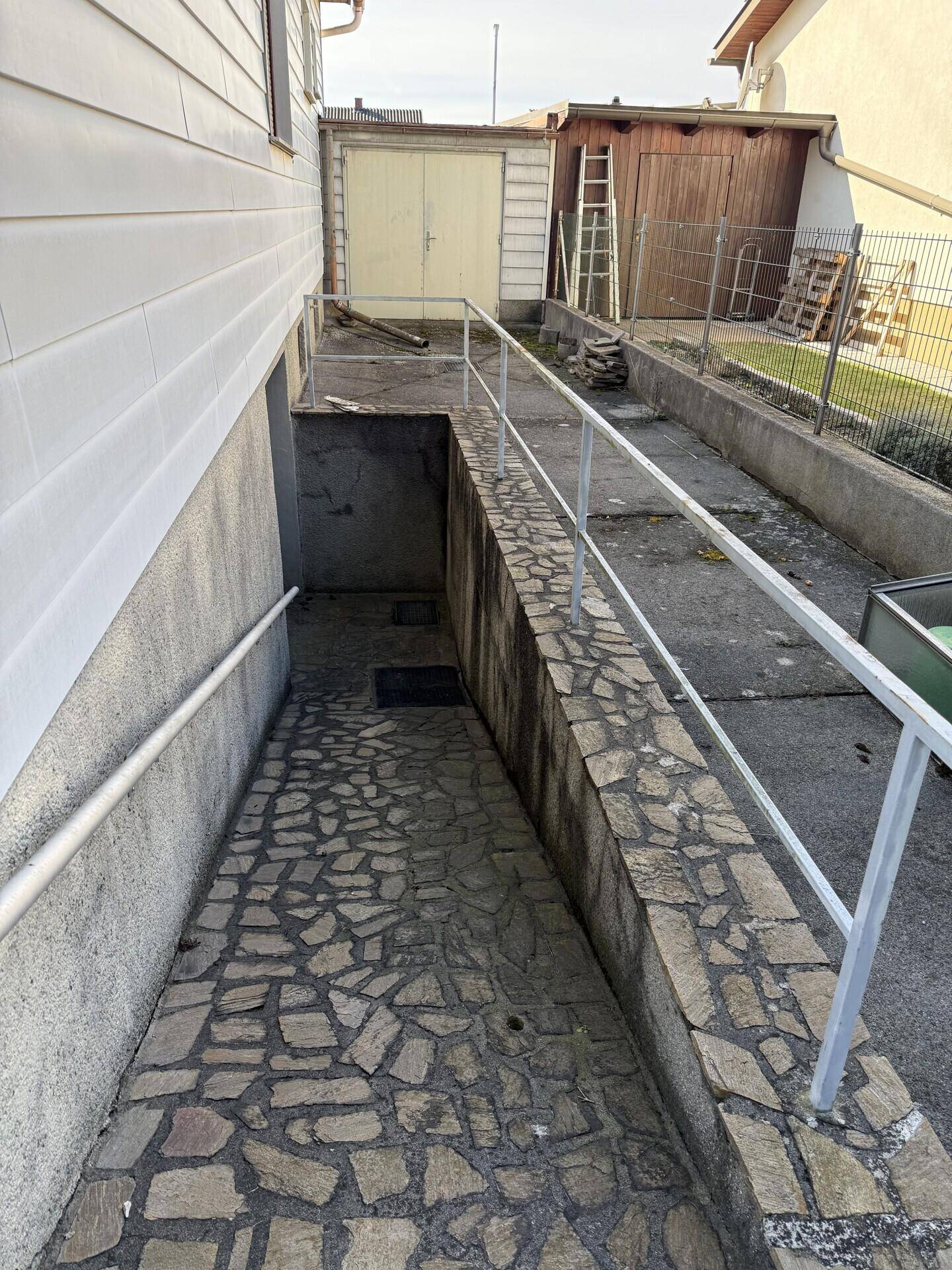 Gepflasterte Rampe mit Handlauf, die zum Untergeschoss führt, umgeben von Steinmauern.