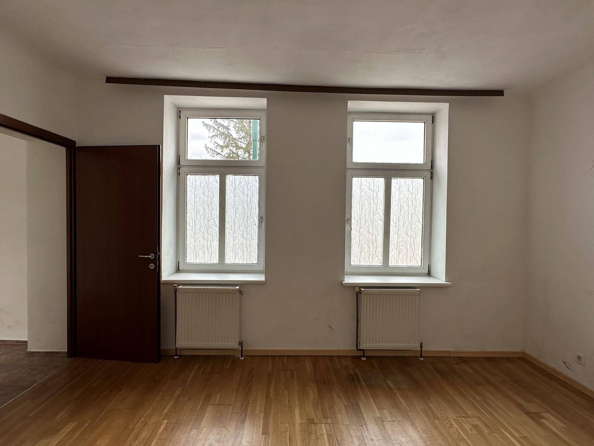 Helles Schlafzimmer mit zwei Fenstern und Holzboden, das Renovierungsbedarf zeigt.