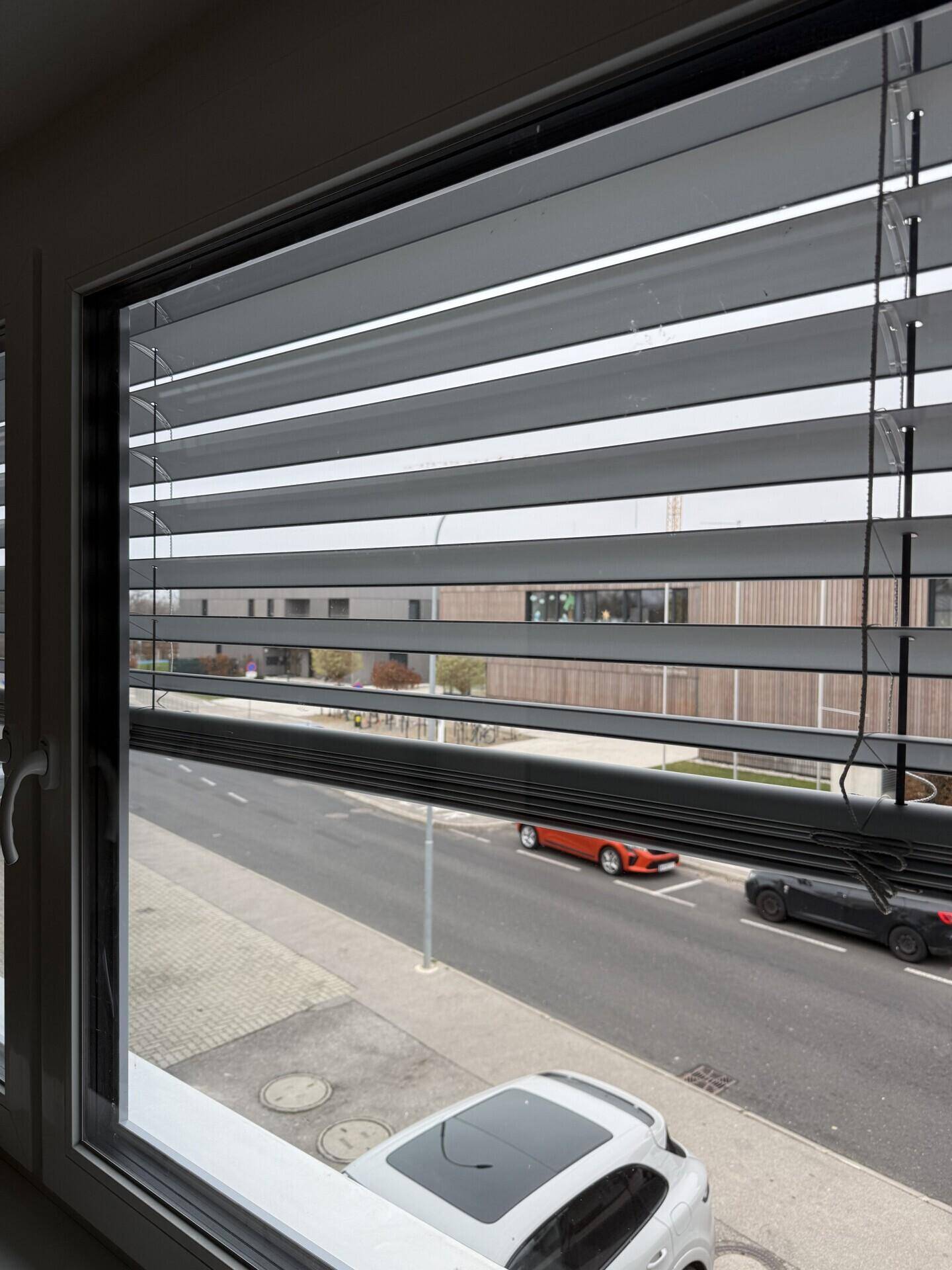 Blick aus einem Fenster mit Jalousien auf eine belebte Straße und gegenüberliegende Gebäude.