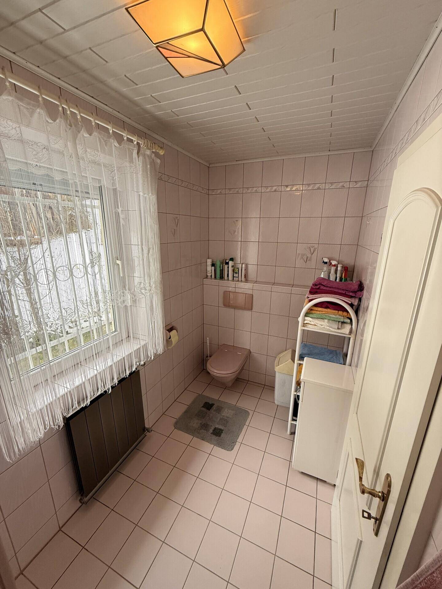 Badezimmer mit rosa Fliesen, Toilette und kleinem Regal in originalem Zustand.