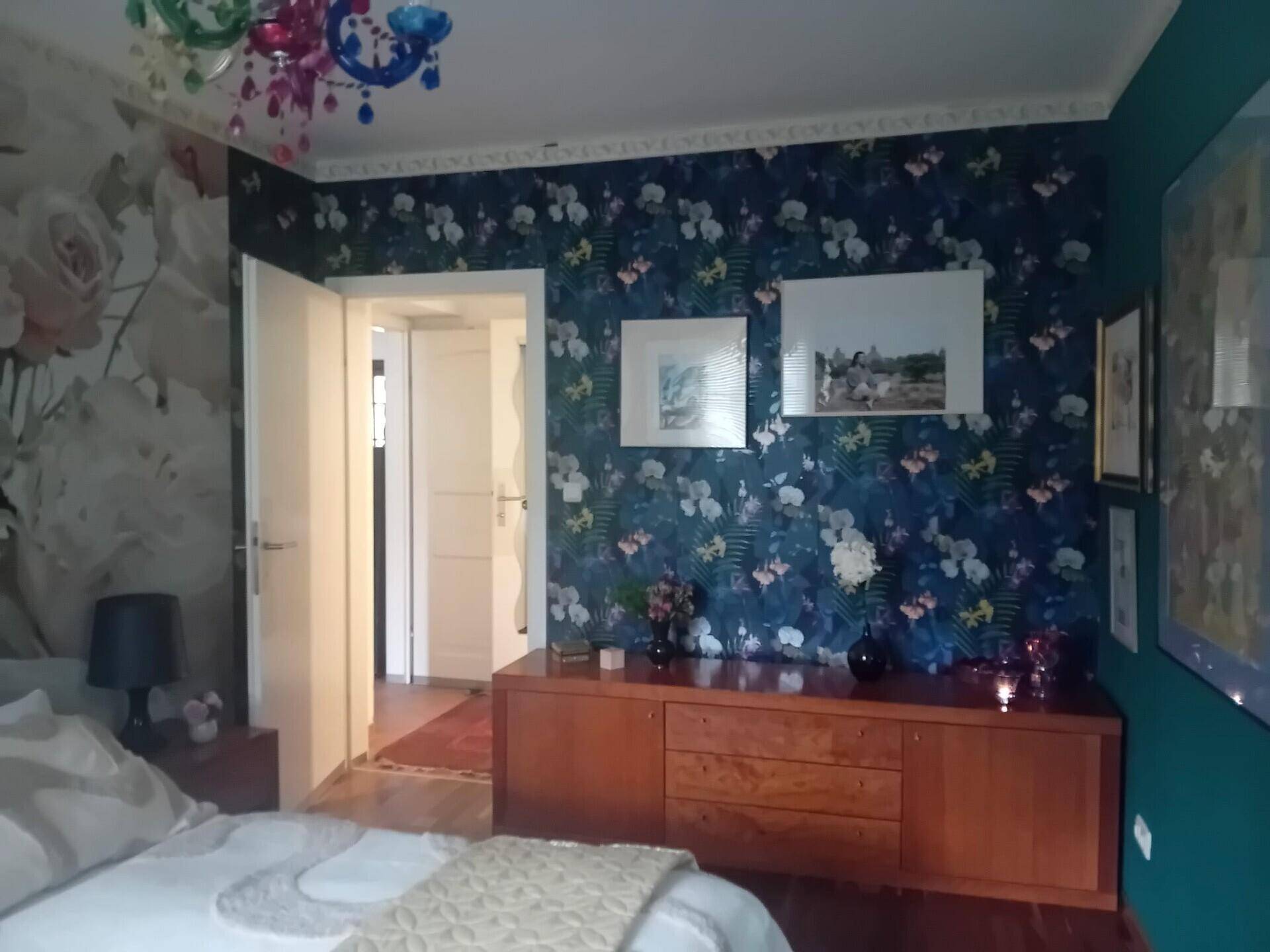 Schlafzimmer mit auffälliger floraler Tapete und Holzkommode, bunte Beleuchtung.