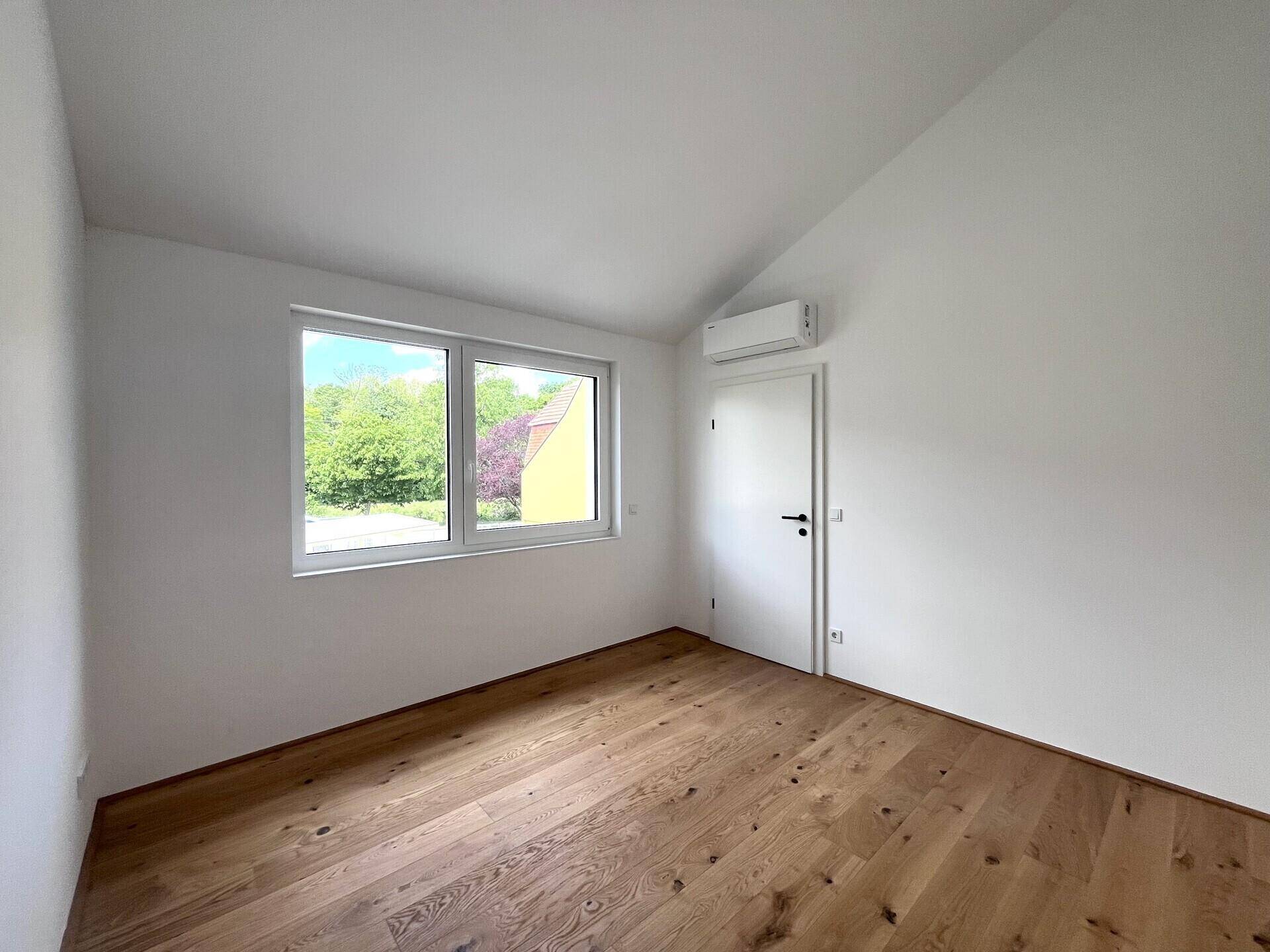 Helles Schlafzimmer mit Holzboden, Dachschräge, Klimaanlage und Fenster mit Blick ins Grüne.