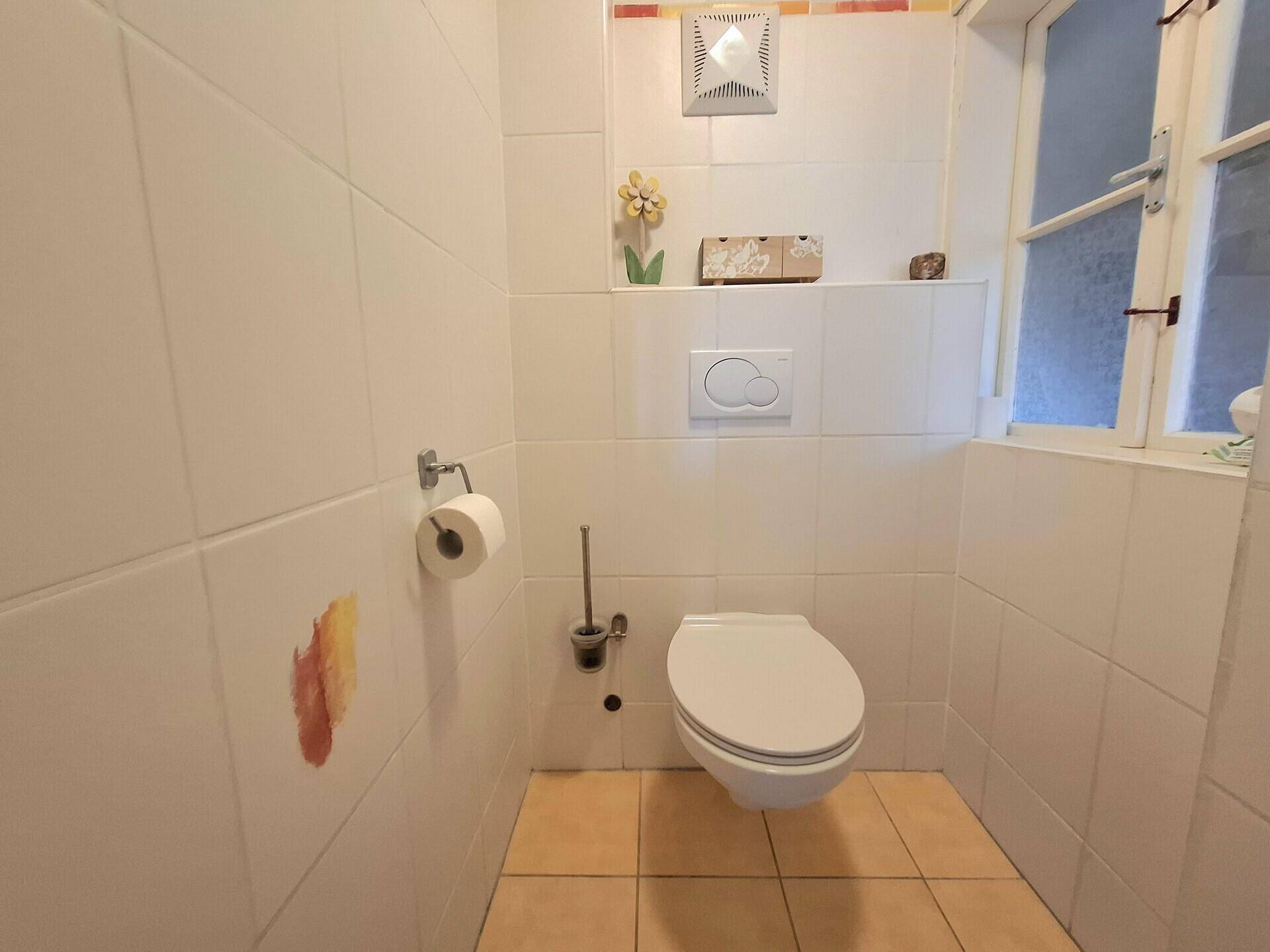 Separate Toilette mit Wand-WC, Fenster und hellen Fliesen, inklusive Lüftungsanlage.