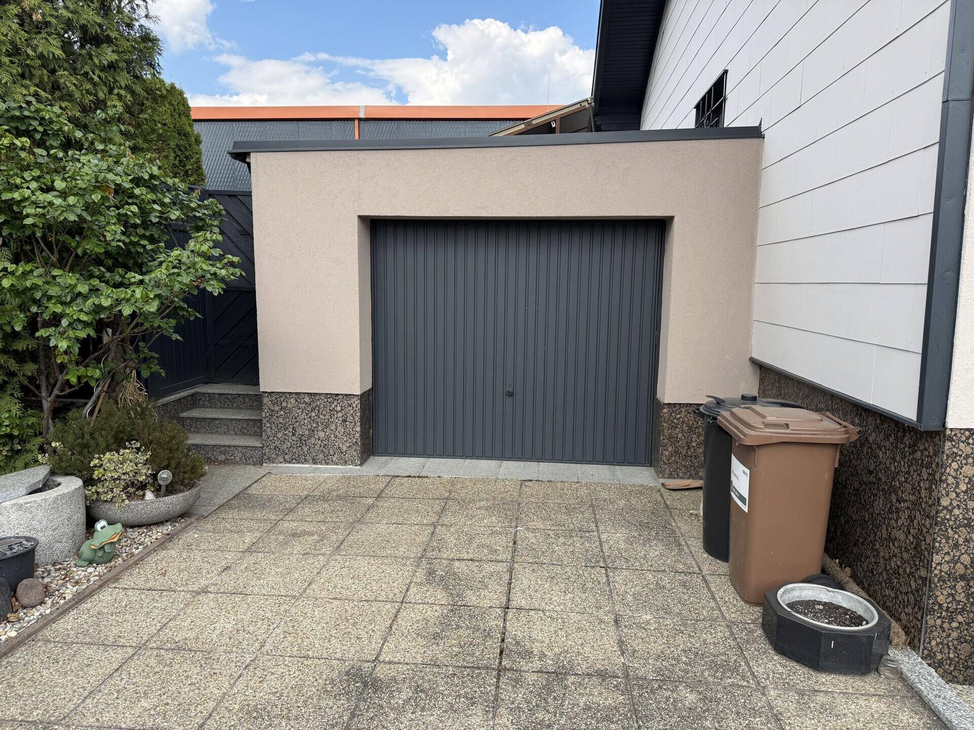 Einzelgarage mit dunklem Tor und gepflastertem Vorplatz neben dem Haus.