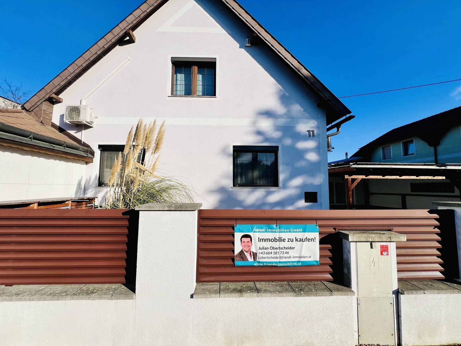 Nahaufnahme der Hausfront mit weißer Fassade, braunem Zaun und einem Immobilien-Werbeschild.