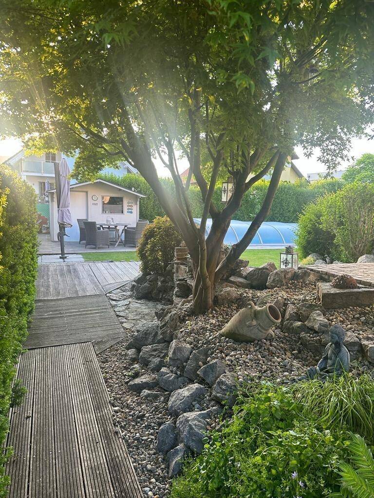 Malerischer Gartenbereich mit altem Baum, Felsen und Holzstegen, führt zu einem Gartenhaus.