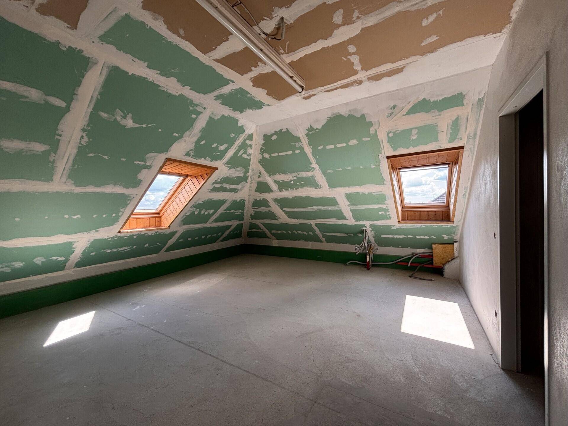 Dachgeschossraum im Rohbau mit schrägen Wänden, Dachfenstern und viel Tageslicht.