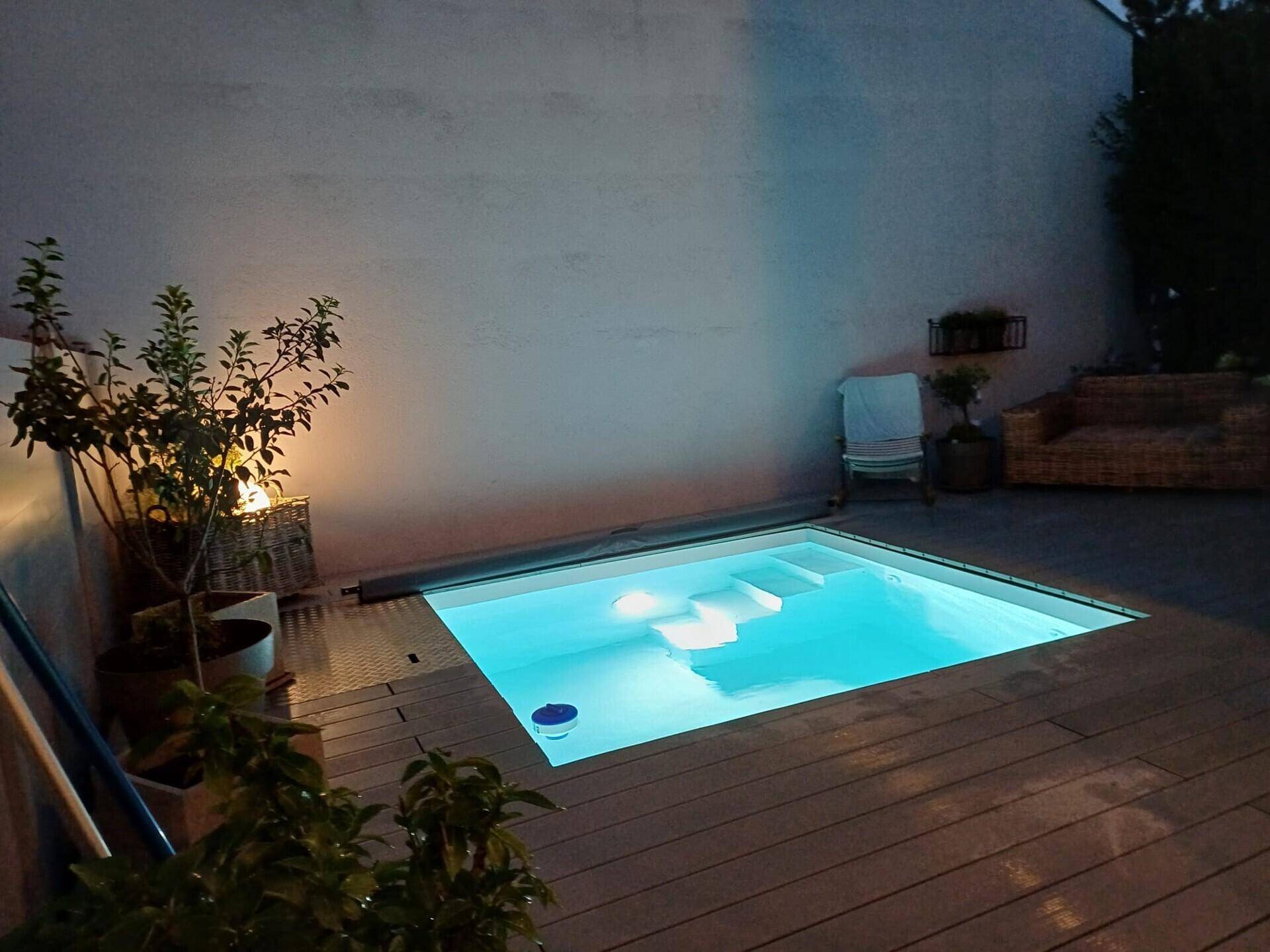 Beleuchteter Poolbereich bei Nacht, umgeben von einem Holzdeck und Pflanzen.
