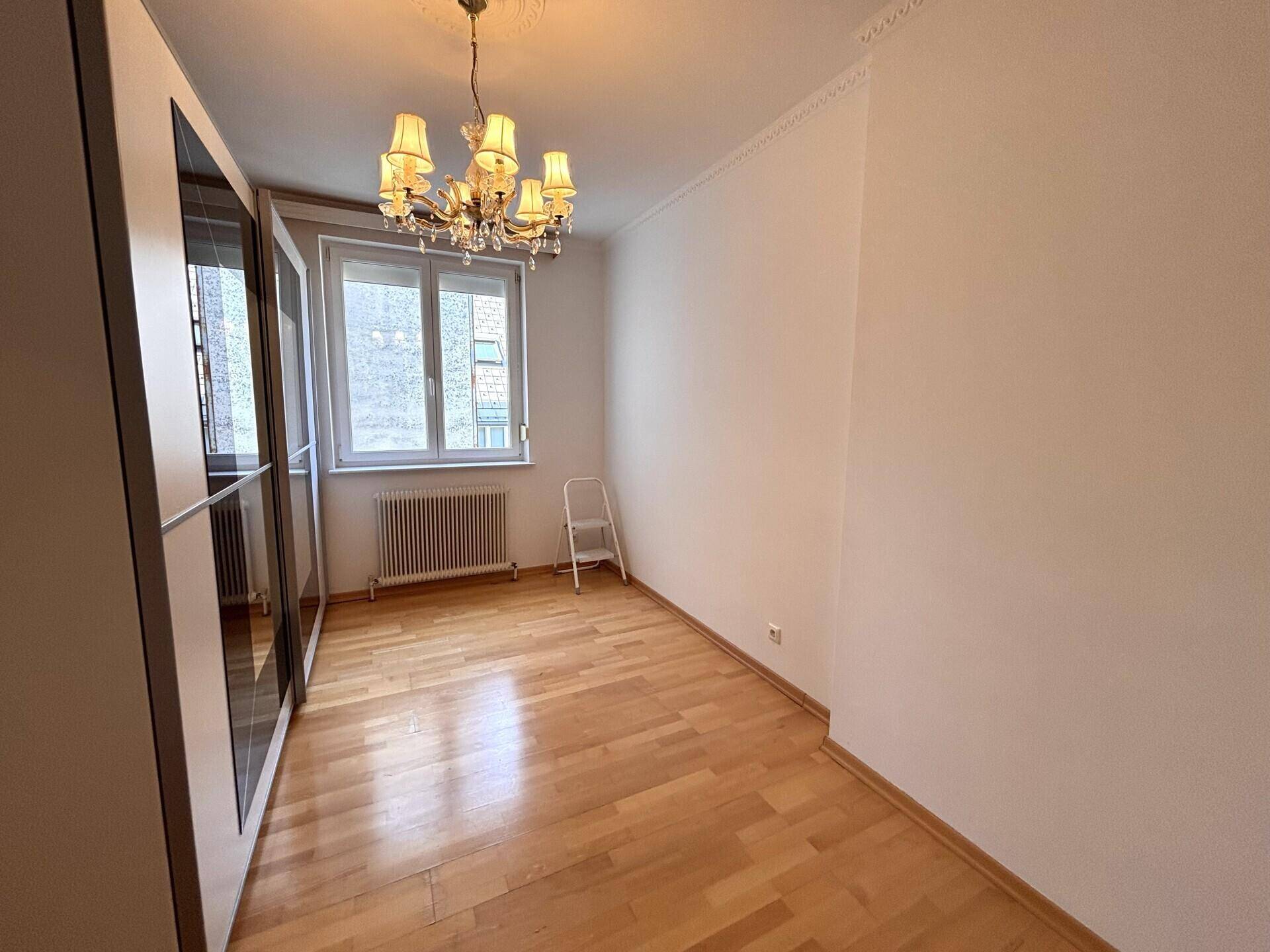 Schlafzimmer mit hellem Holzboden, Spiegelschrank und Fenster, das Tageslicht hereinlässt.