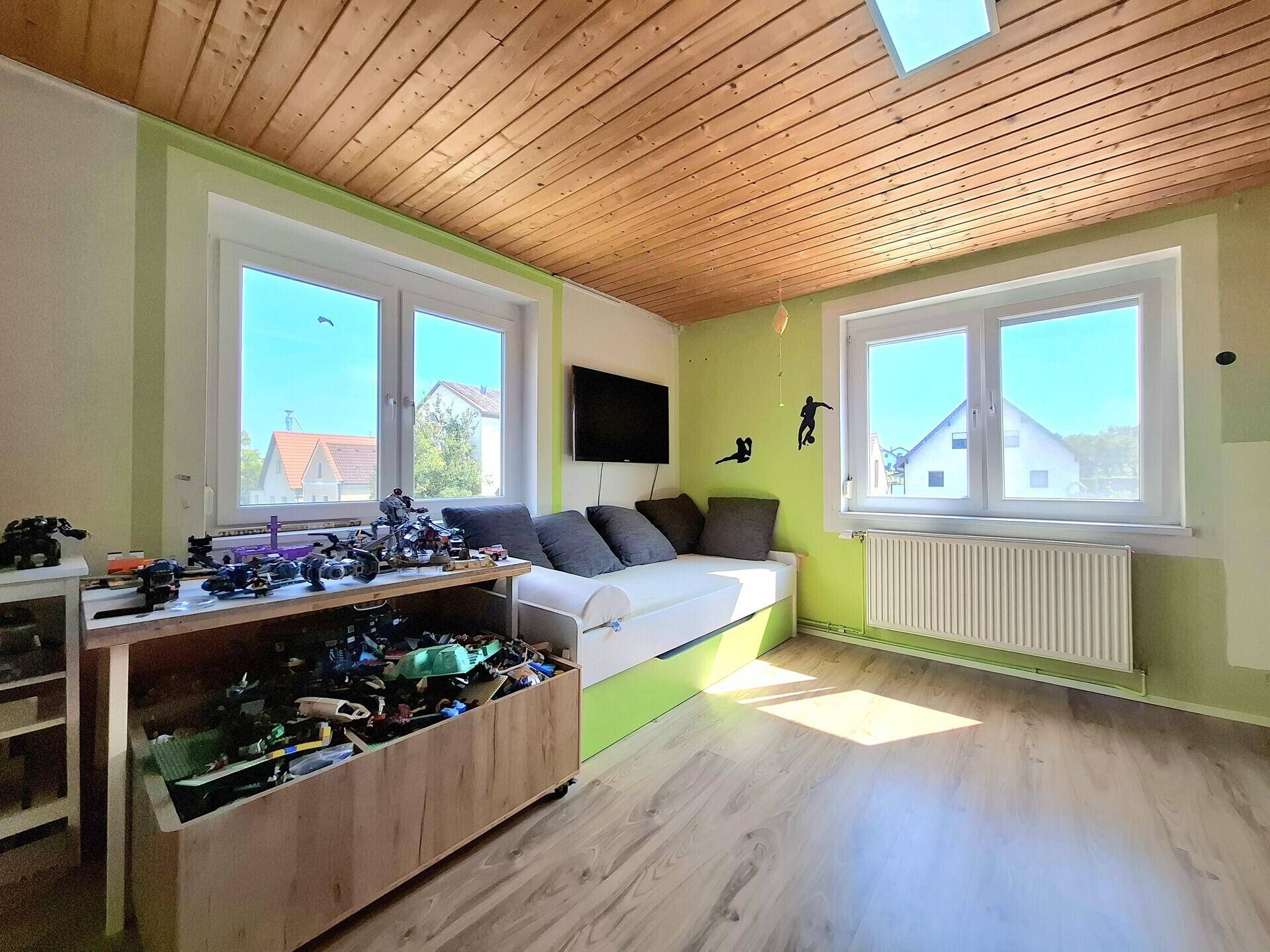 Kinderzimmer mit Holzbalkendecke, Spielzeug und hellgrünen Wänden, gut beleuchtet durch Fenster.