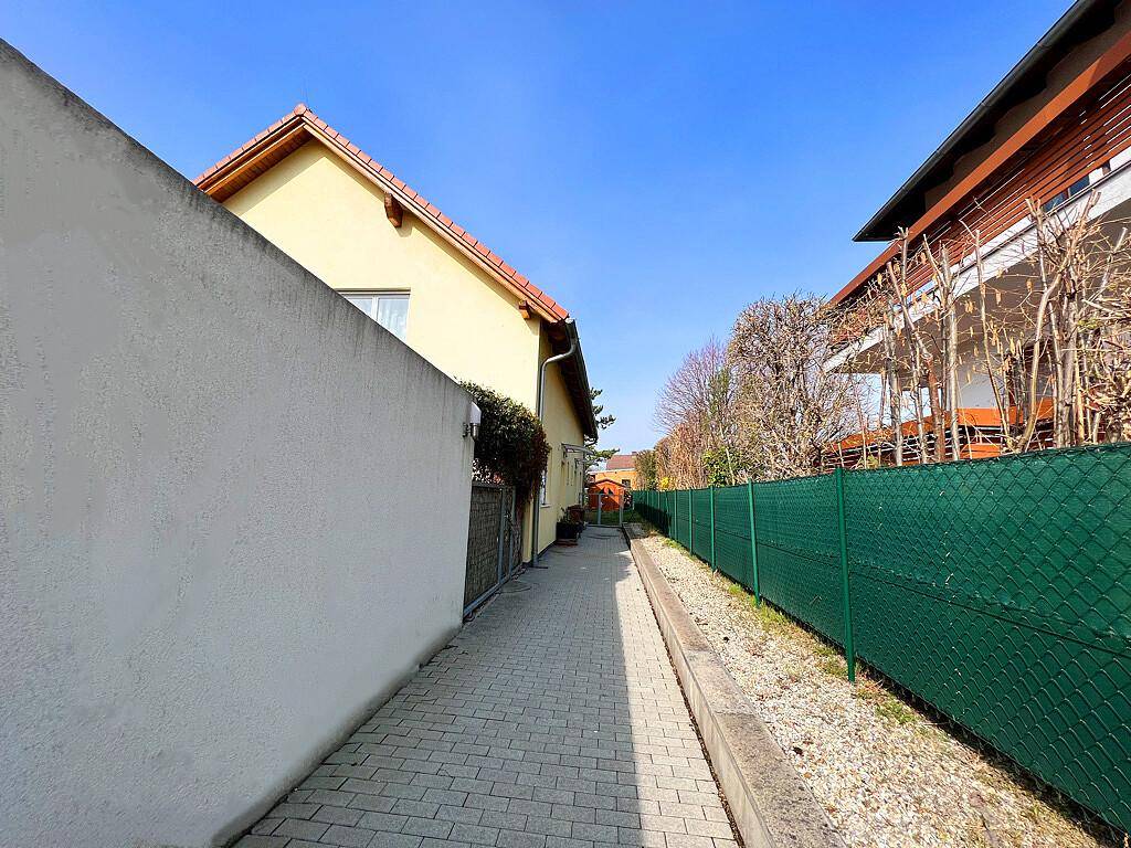 Schmaler gepflasterter Weg zwischen zwei Häusern, begrenzt von einer Mauer und einem grünen Zaun.