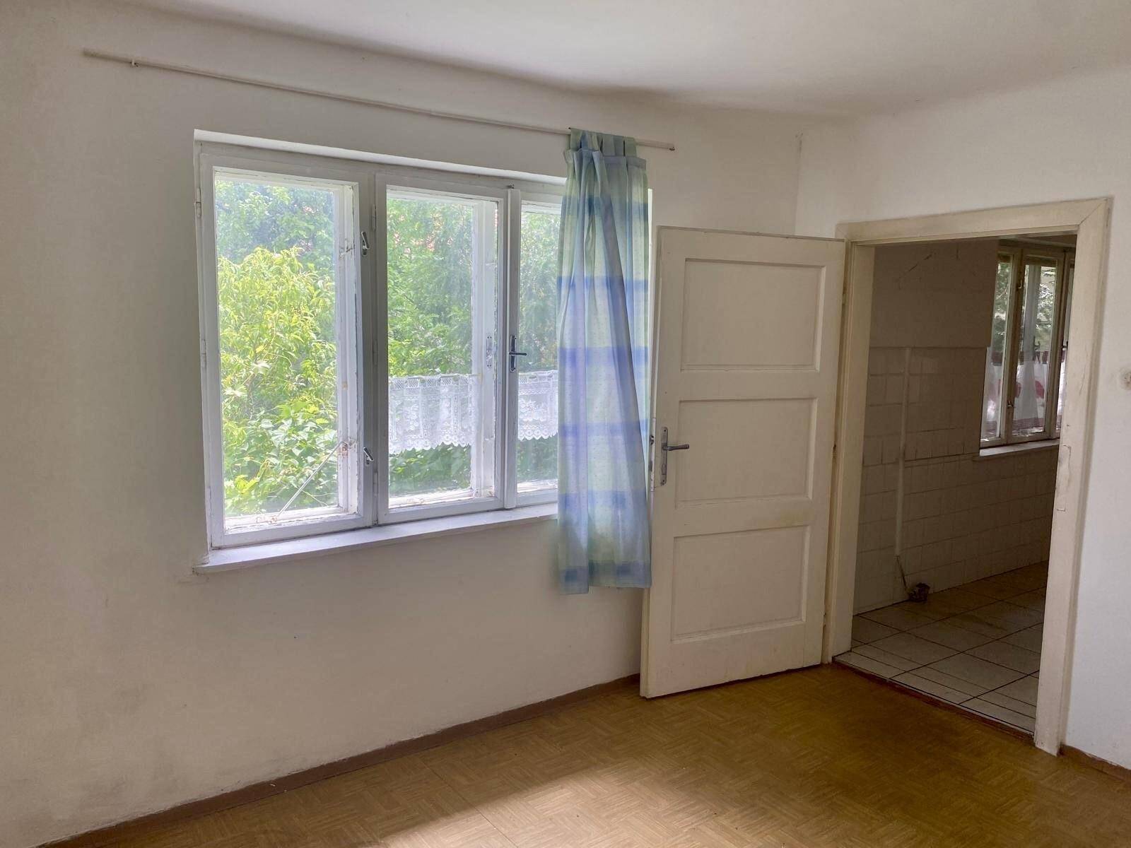 Helles Zimmer mit Holzboden, Fenster und einer Tür, die zu einem weiteren Raum führt.