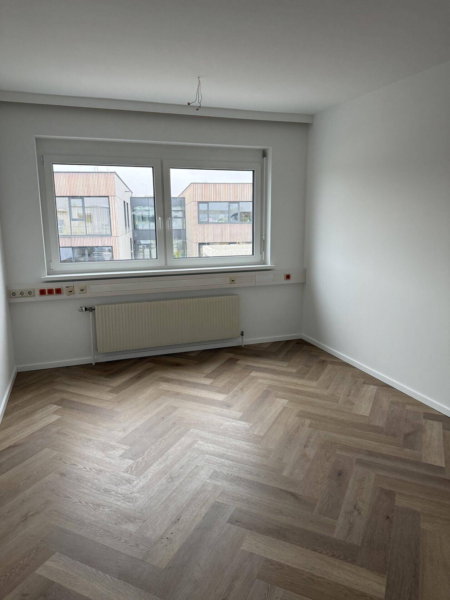 Heller, leerer Büroraum mit Fischgrätparkett, einem Fenster und einem Heizkörper an der Wand.