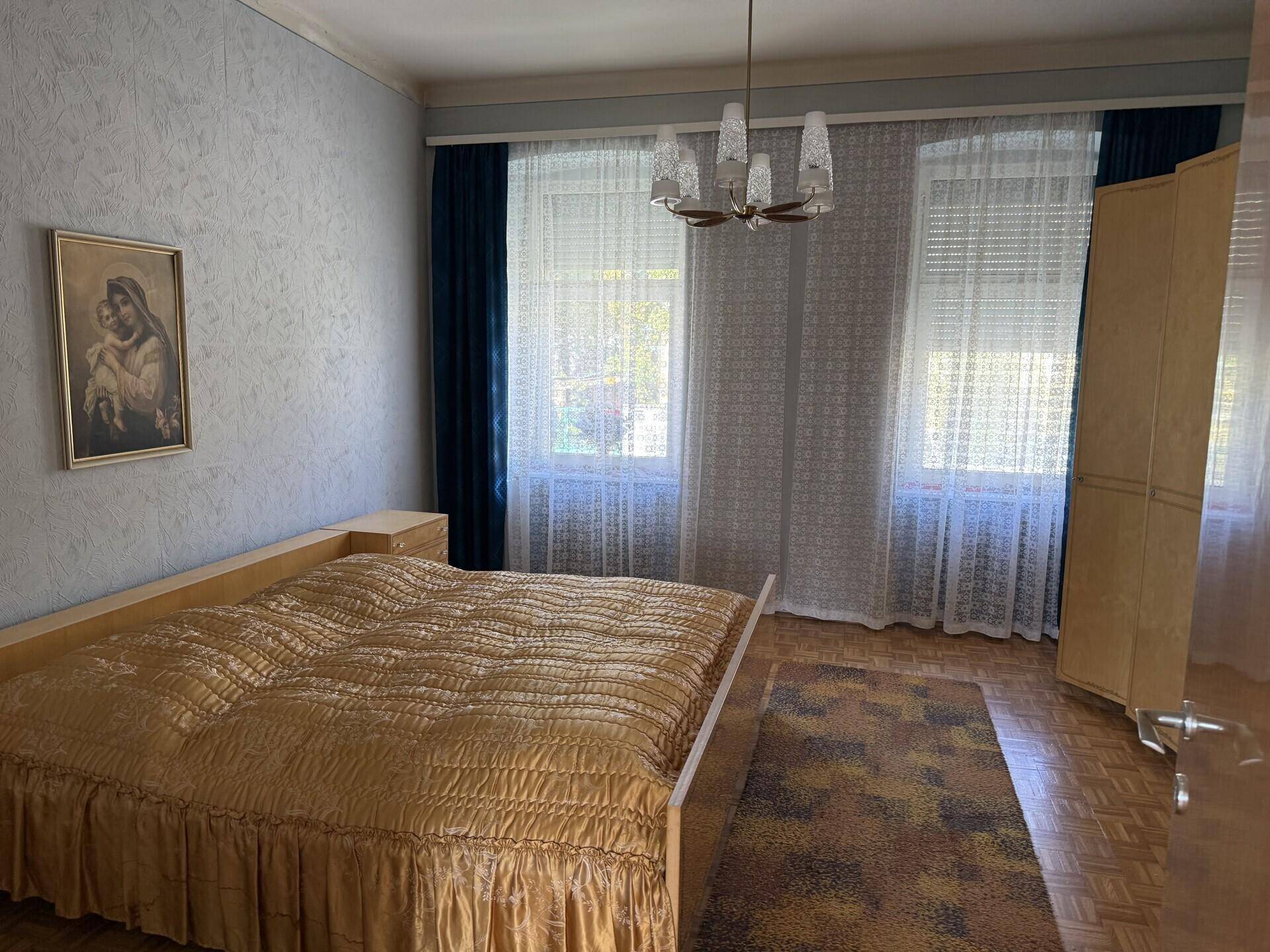 Geräumiges Schlafzimmer mit Doppelbett, Parkettboden und großen Fenstern mit Vorhängen.