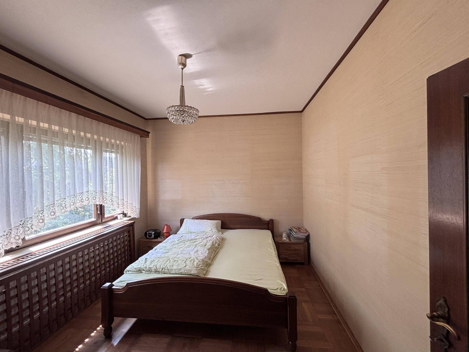 Schlafzimmer mit Holzbett, Parkettboden und großem Fenster mit Vorhängen.