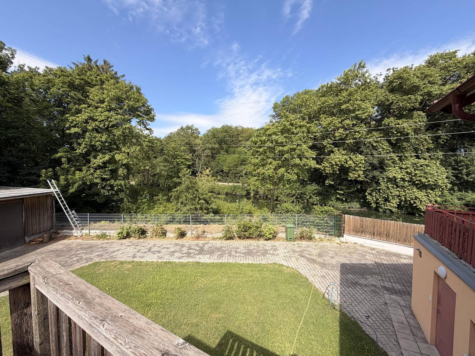 Weitläufiger Blick über den Garten mit Rasenfläche und dichtem Baumbestand im Hintergrund.
