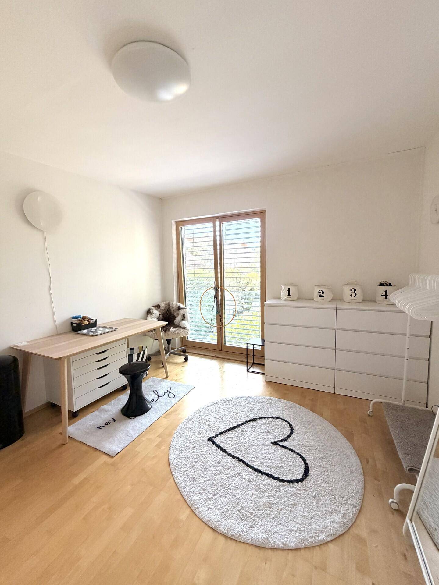 Helles Zimmer mit Holzboden, Schreibtisch und großem Fenster mit Jalousien.