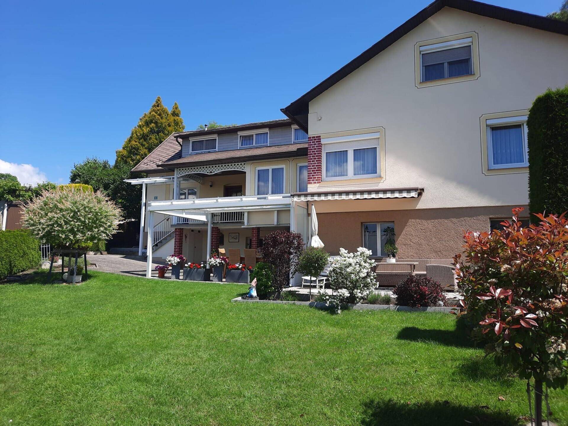 Hausfassade mit großem Garten, überdachter Terrasse und Balkon im Obergeschoss.