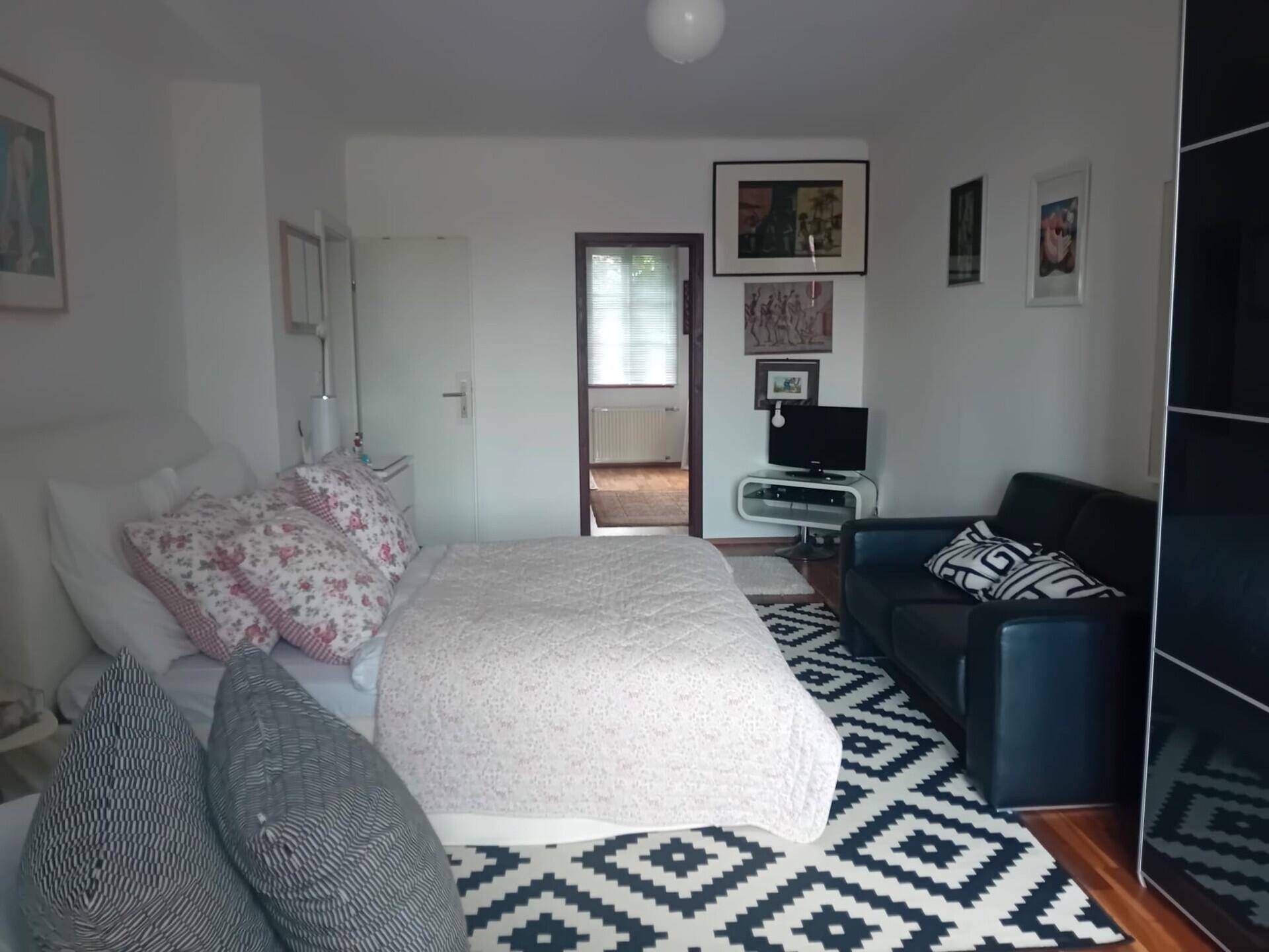 Gemütliches Schlafzimmer mit Bett, kleinem Sofa und Fernseher, Holzboden.