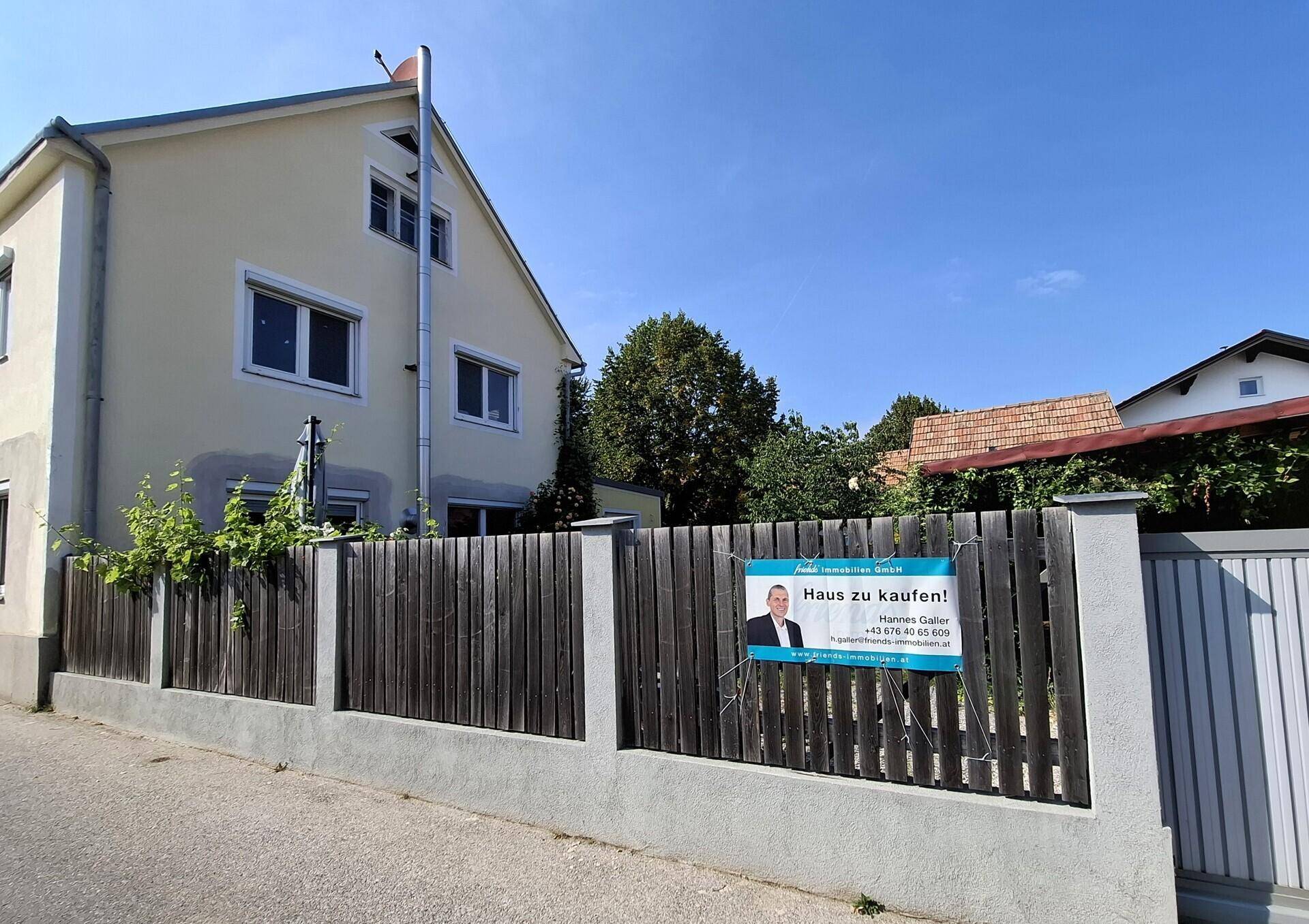 Ansicht des Hauses von der Straße mit Zaun und einem "Haus zu kaufen" Schild.