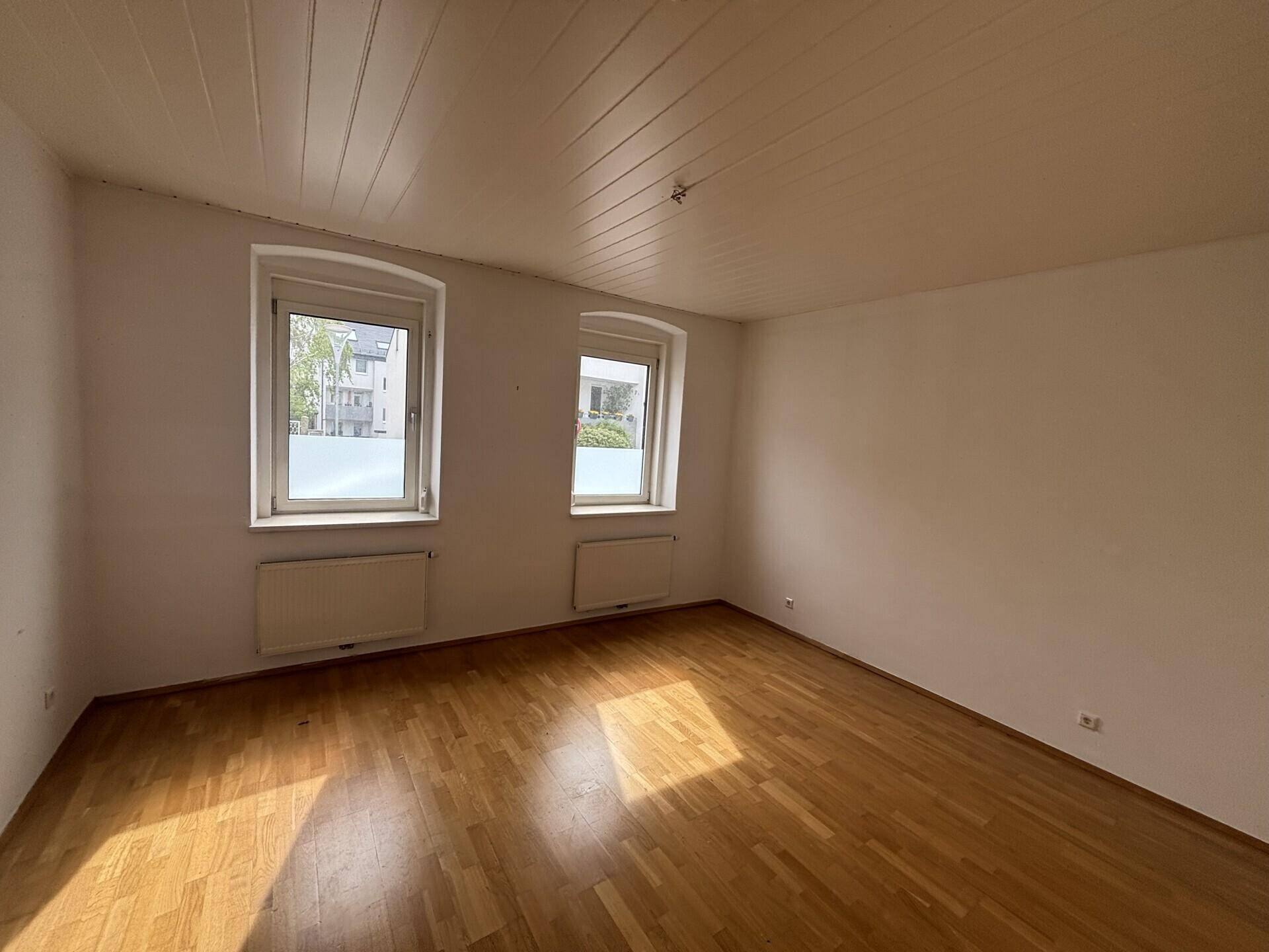 Schlafzimmer mit hellem Parkettboden, zwei Fenstern und Holzdeckenverkleidung.