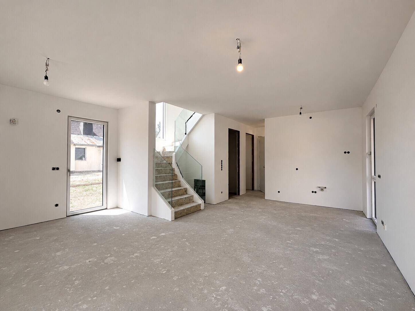 Geräumiger, unfertiger Wohnbereich mit offener Treppe und Betonboden, wartet auf den Innenausbau.