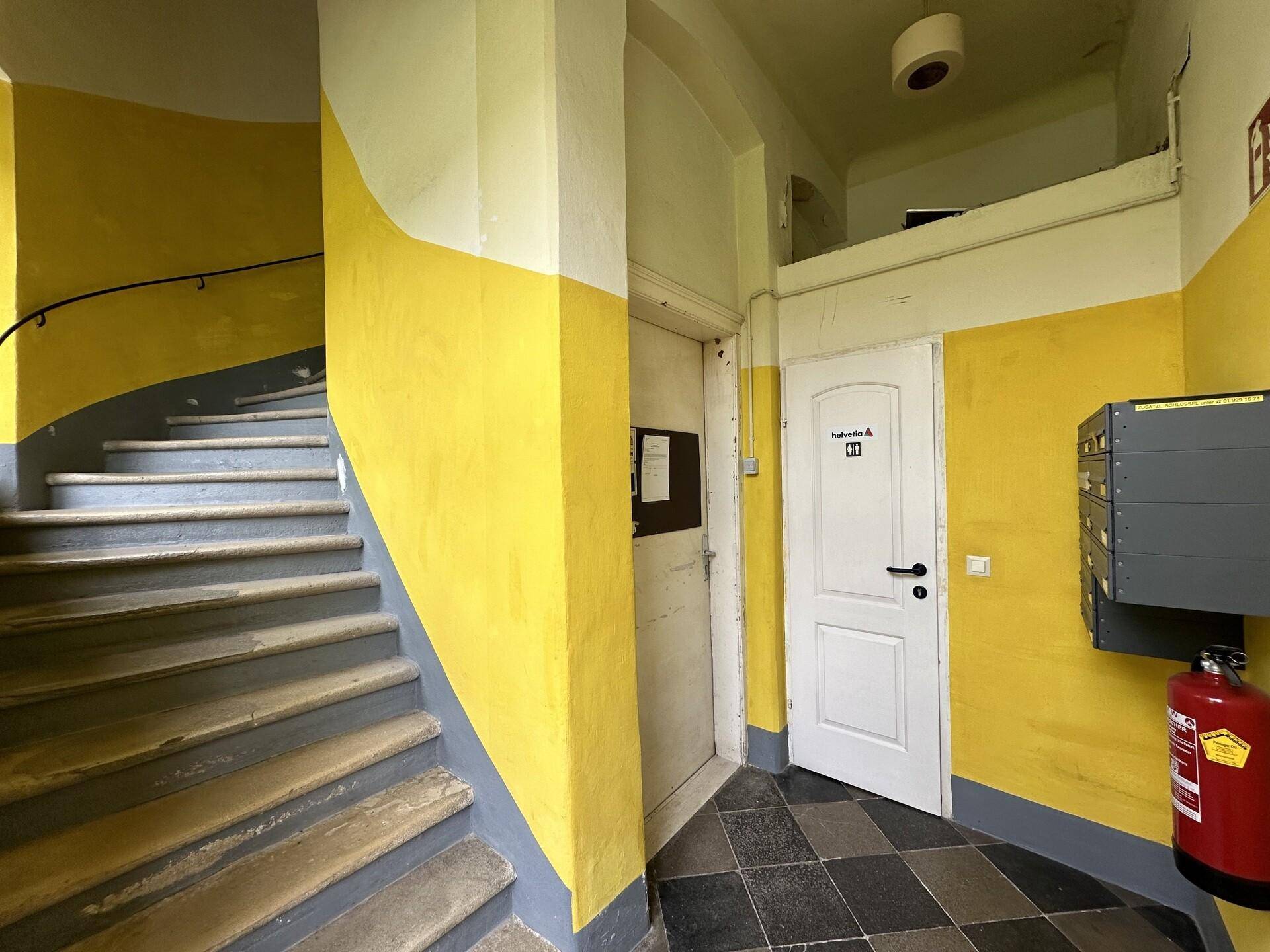 Treppenhaus mit gelb gestrichenen Wänden und dunklem Fliesenboden, mit Briefkästen und Feuerlöscher.
