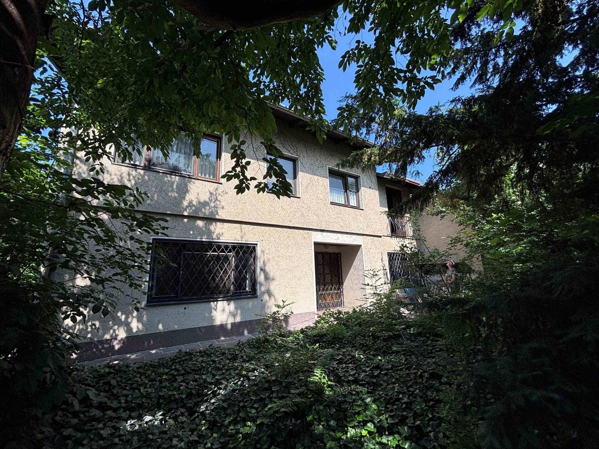 Hausfassade umgeben von dichtem Grün und Bäumen an einem sonnigen Tag.
