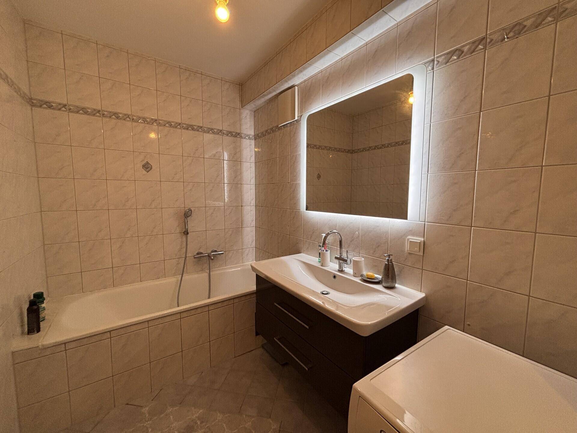Helles Badezimmer mit beigefarbenen Fliesen, Badewanne und modernem Waschtisch mit beleuchtetem Spiegel.