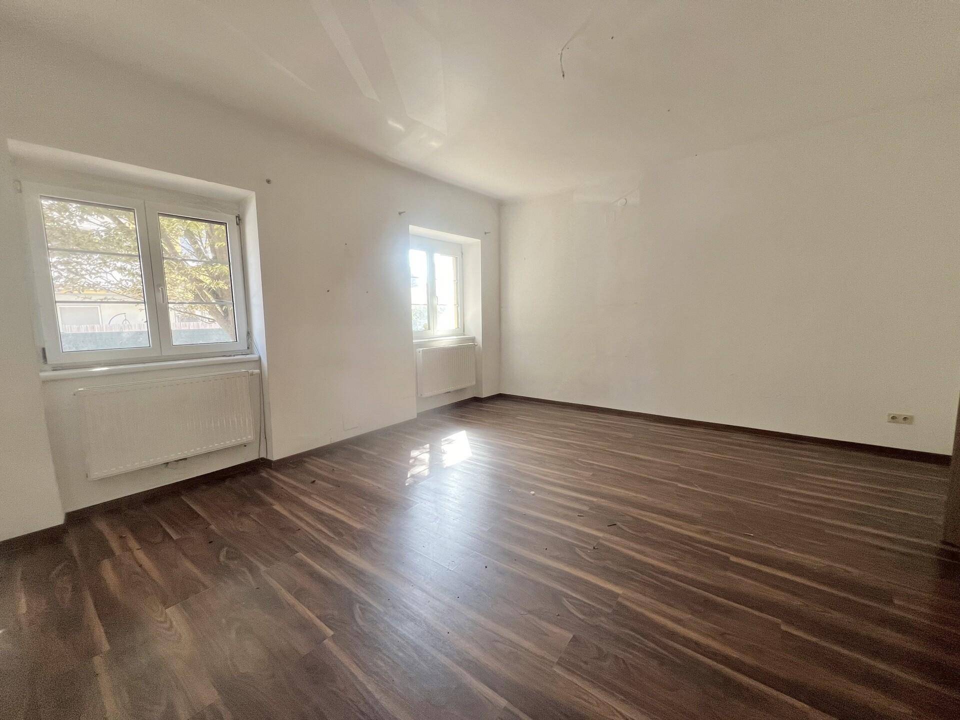 Geräumiger, heller Raum mit dunklem Laminatboden und zwei Fenstern.