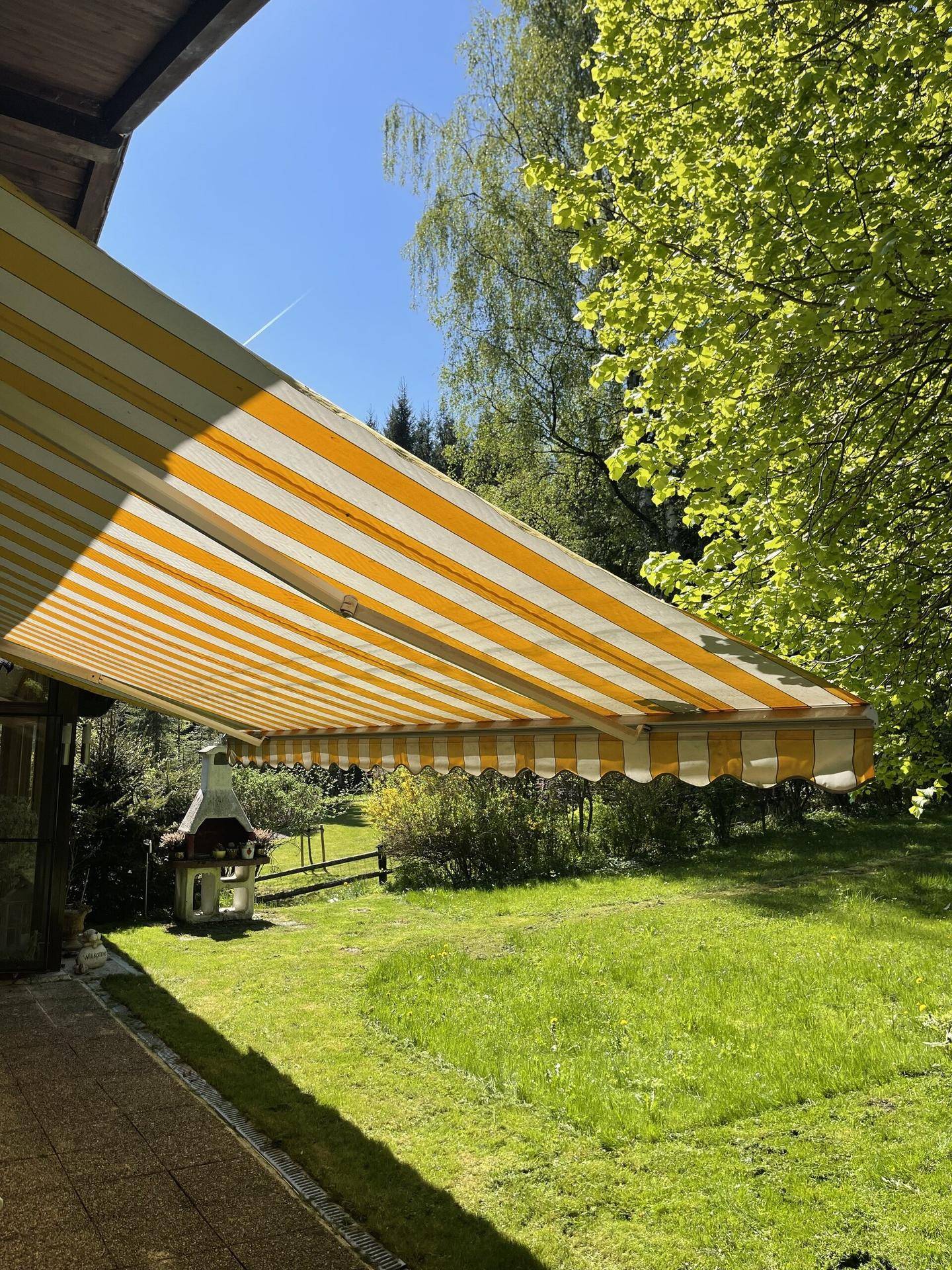 Überdachte Terrasse mit ausgefahrener gelb-weiß gestreifter Markise und Grillplatz.