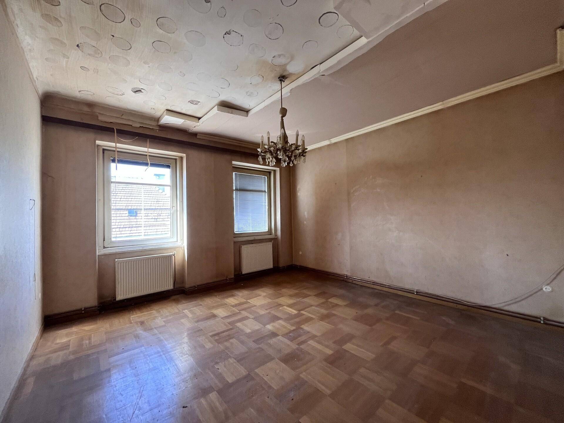 Großes Zimmer mit Fischgrätparkett, Kronleuchter und zwei Fenstern, renovierungsbedürftig.