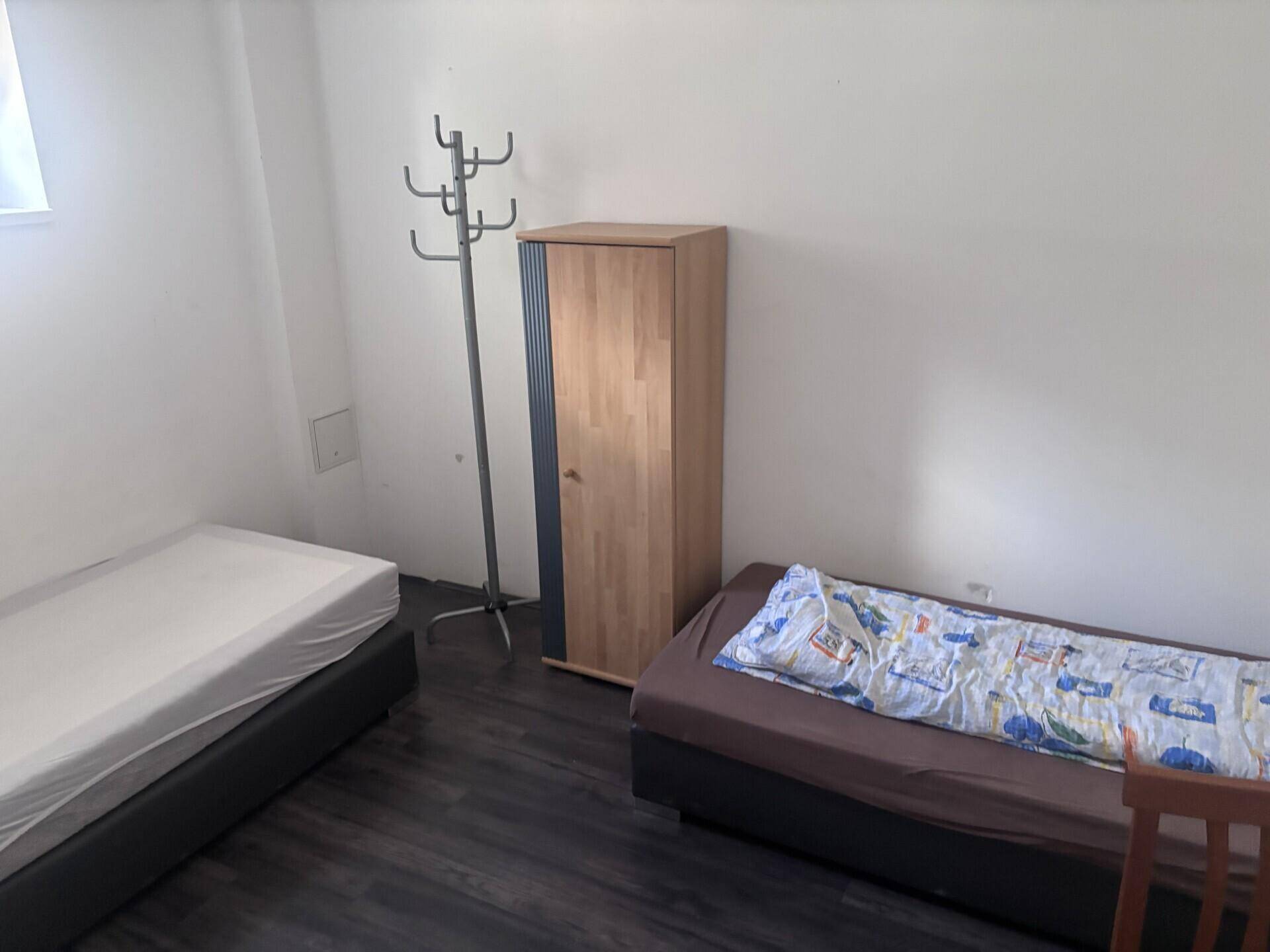Schlafzimmer mit zwei Betten, einem Kleiderschrank und dunklem Holzboden in schlichtem Design.