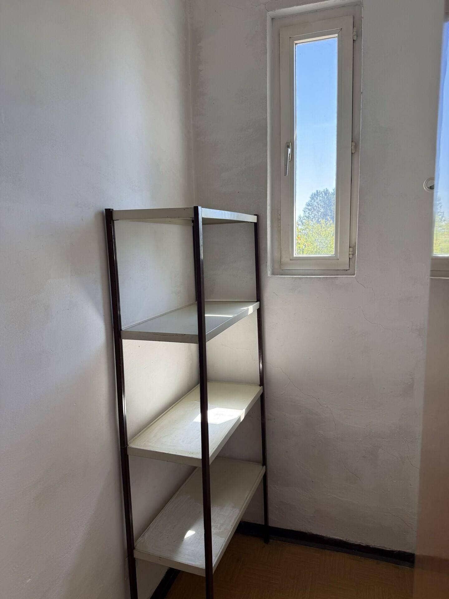 Einfacher Abstellraum mit einem Regal und einem hohen Fenster, das Tageslicht hereinlässt.