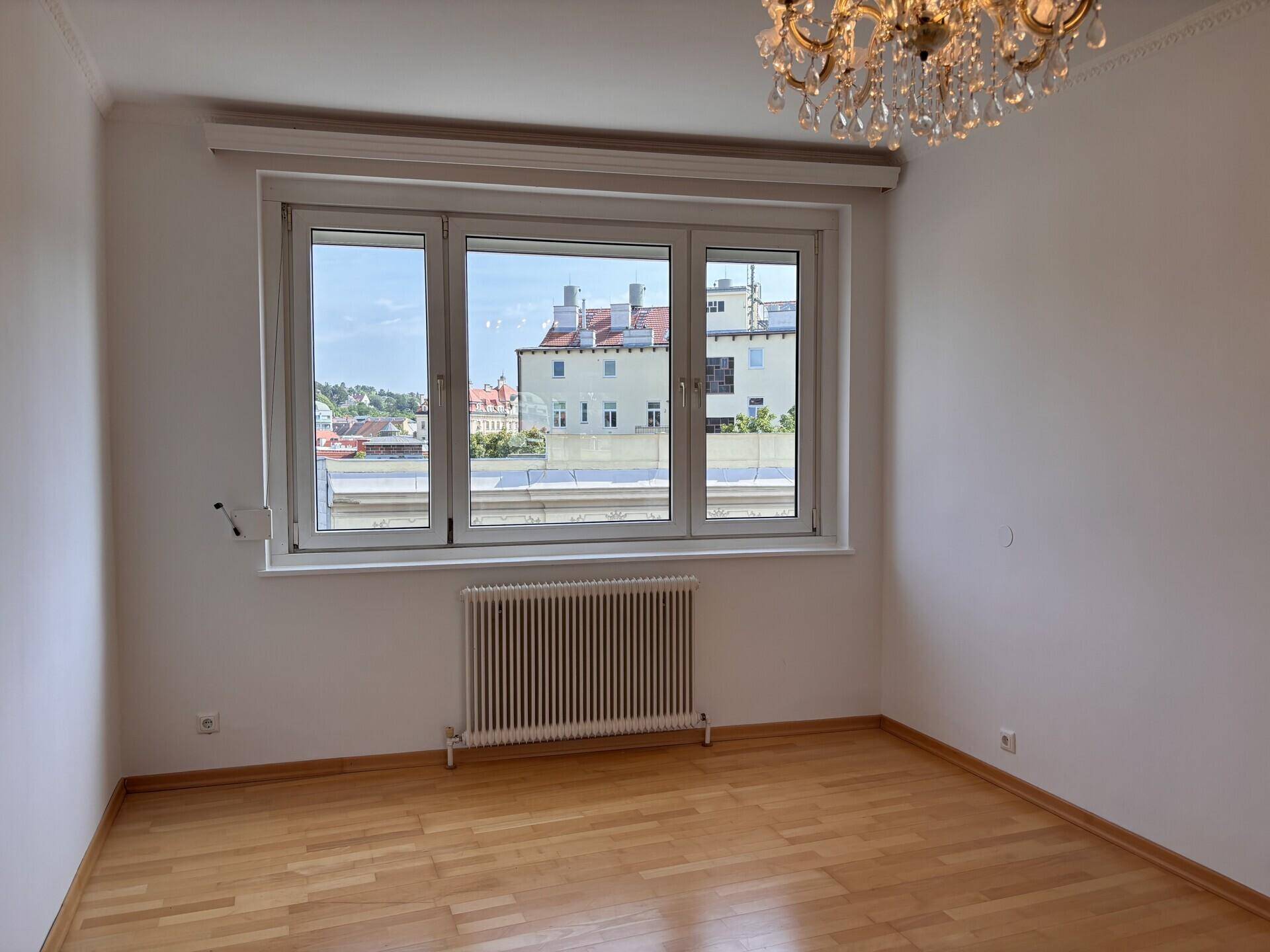 Helles Zimmer mit Holzboden und Fenster mit Blick auf die Dächer und Gebäude der Stadt.