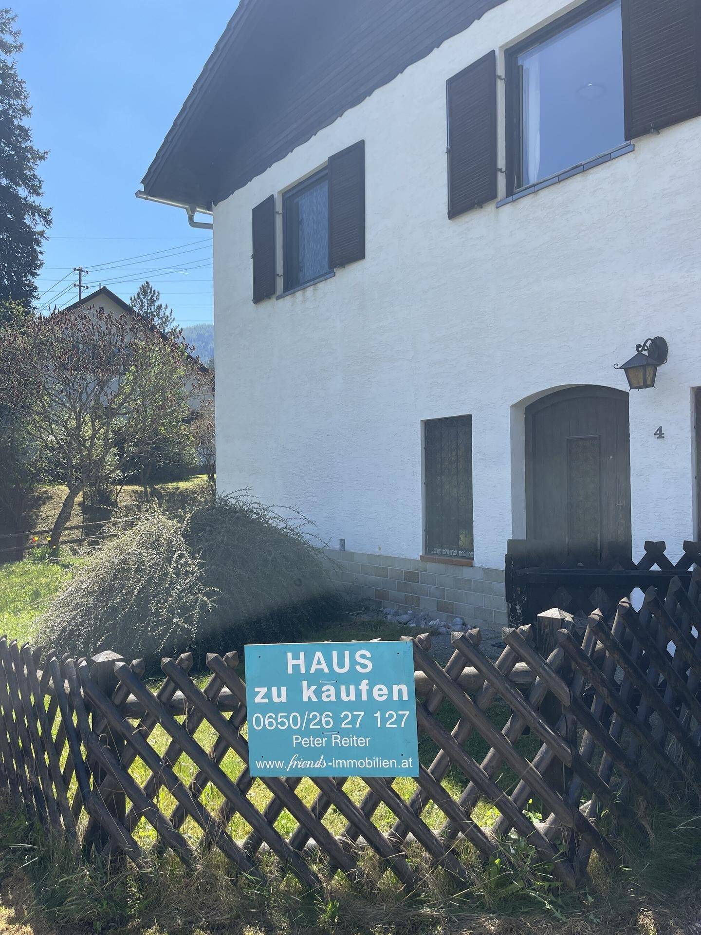 Vorderansicht des Hauses mit einem "Haus zu kaufen" Schild am Holzzaun.