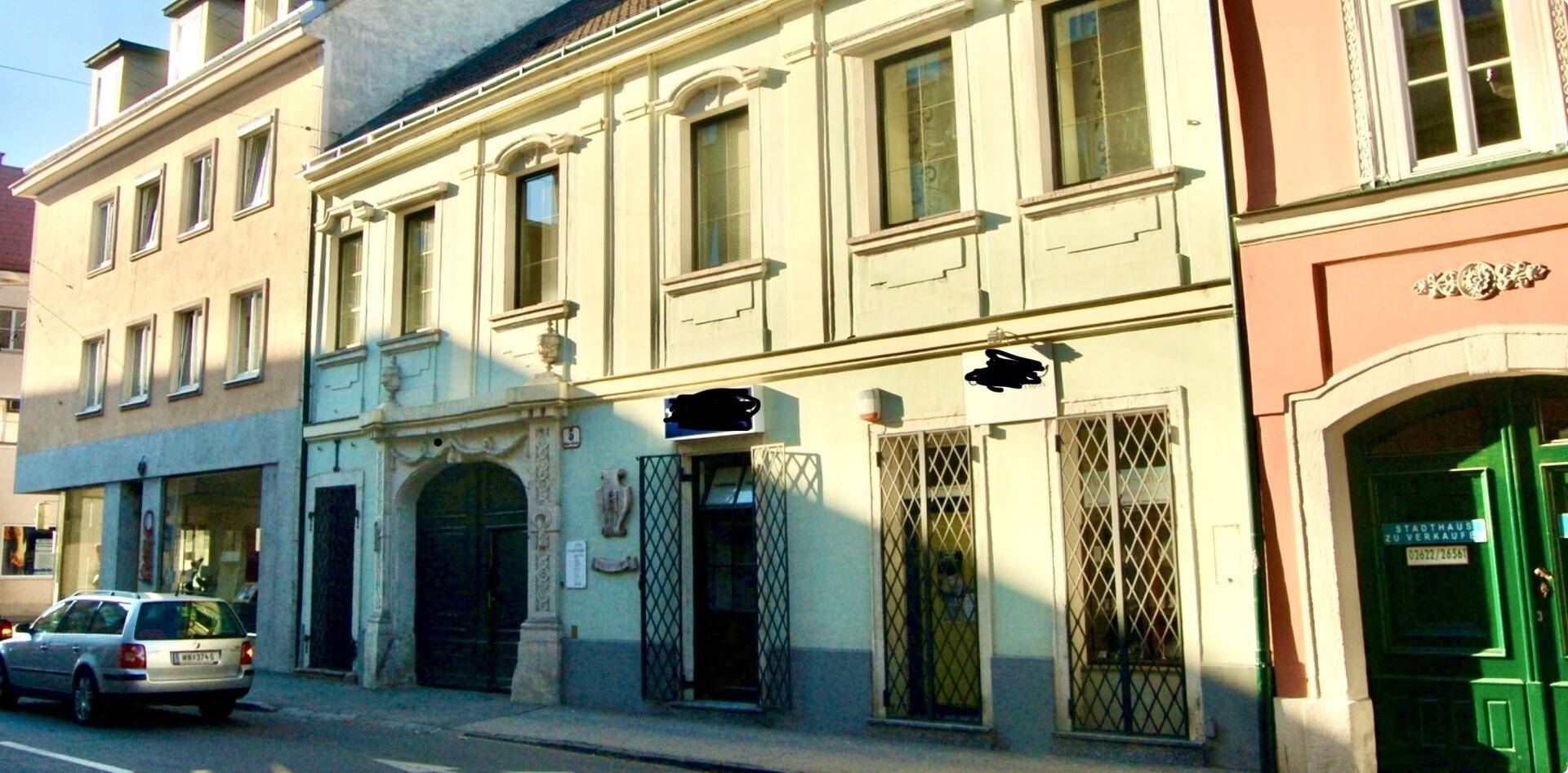 Historisches Gebäude mit grünen Toren und vergitterten Fenstern, umgeben von anderen Stadtgebäuden.