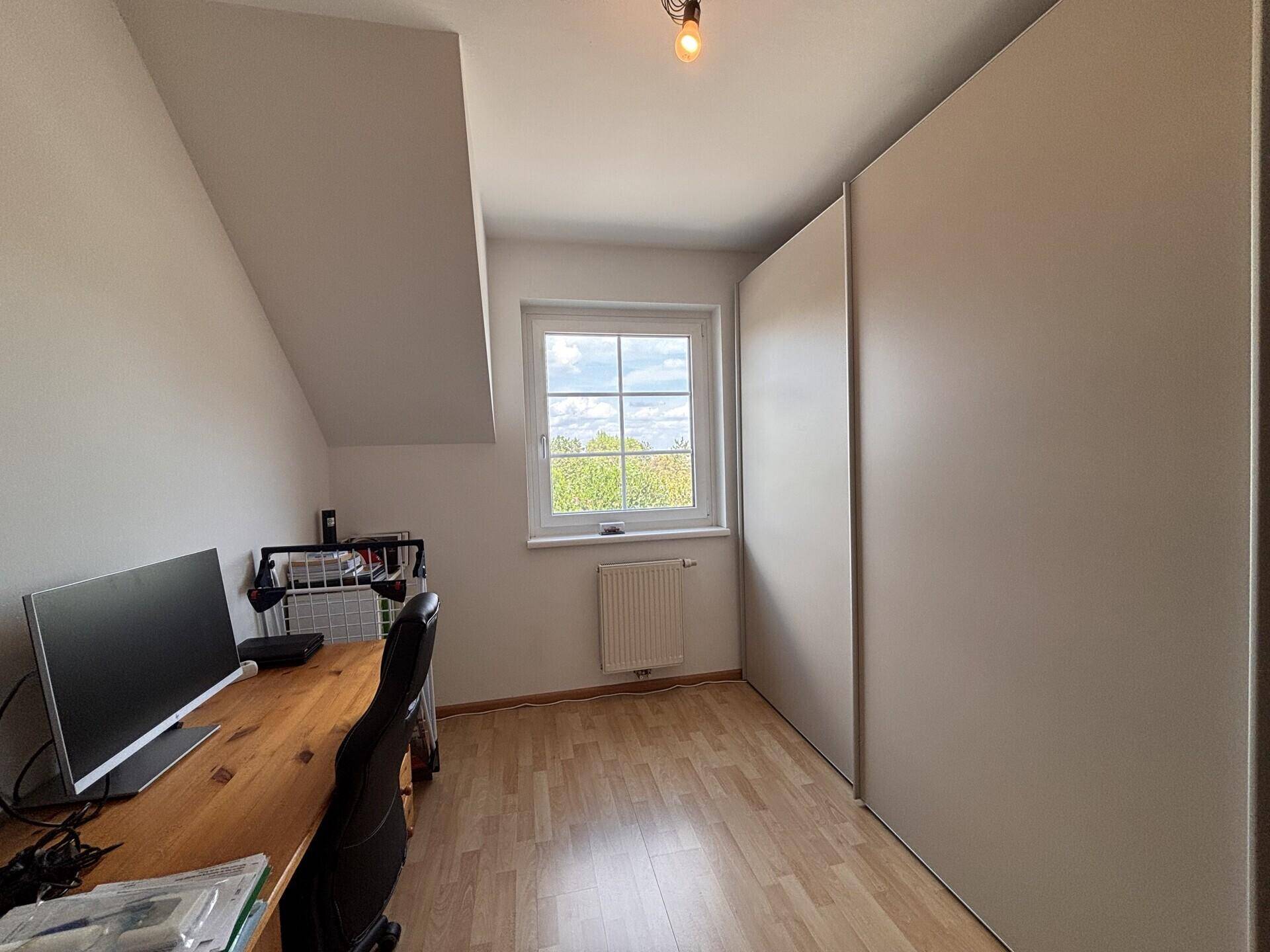 Helles Arbeitszimmer mit Holzschreibtisch, Computer und großem Einbauschrank unter schräger Decke.