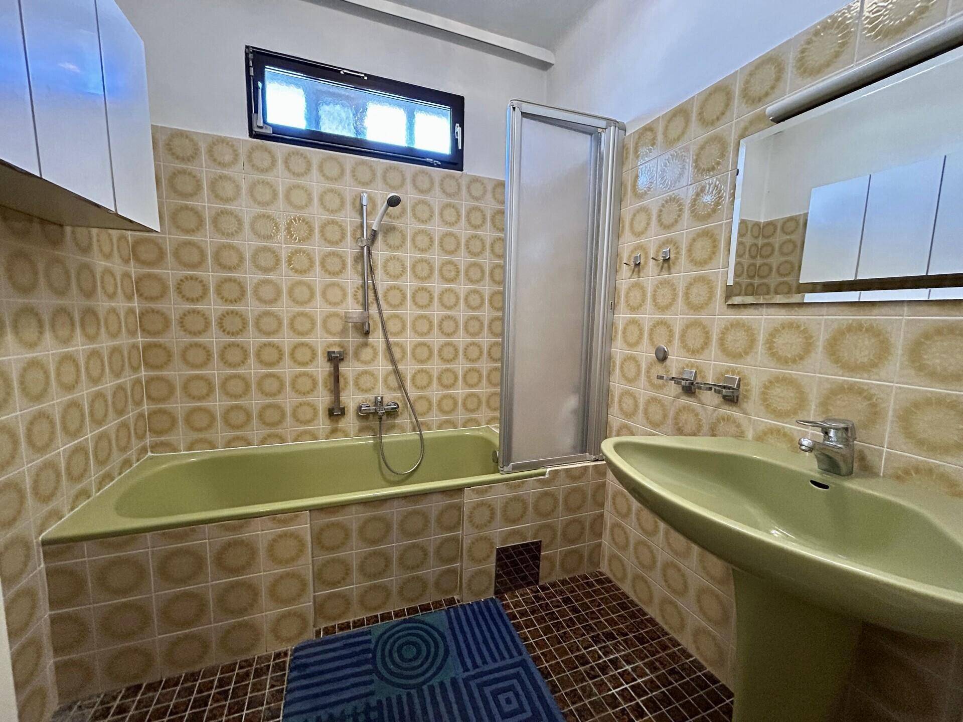 Badezimmer im 80er-Jahre-Stil mit grüner Badewanne, Waschbecken und gemusterten Fliesen.