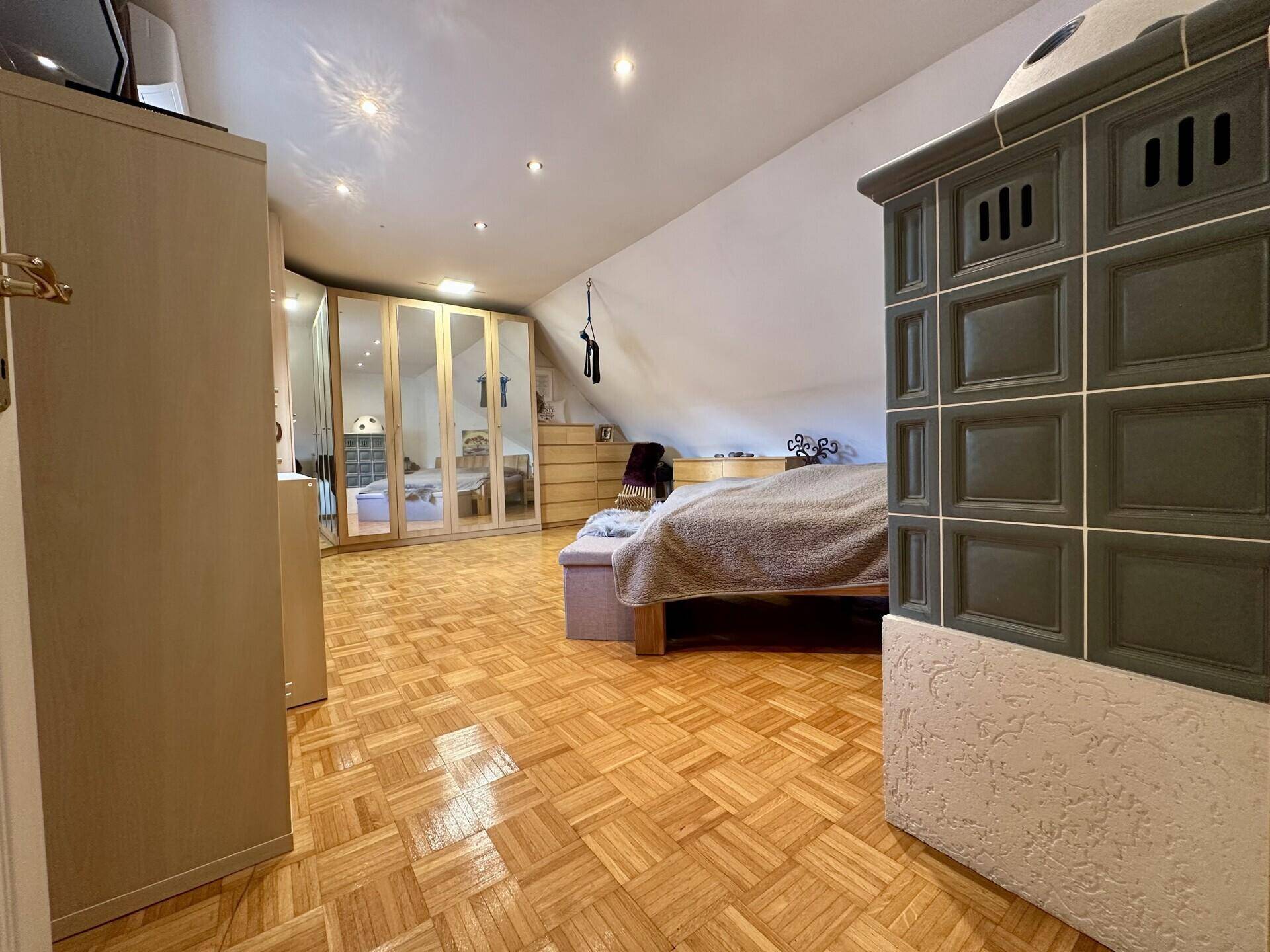 Schlafzimmer im Dachgeschoss mit Parkettboden, Bett, Spiegelschränken und einem Kachelofen.