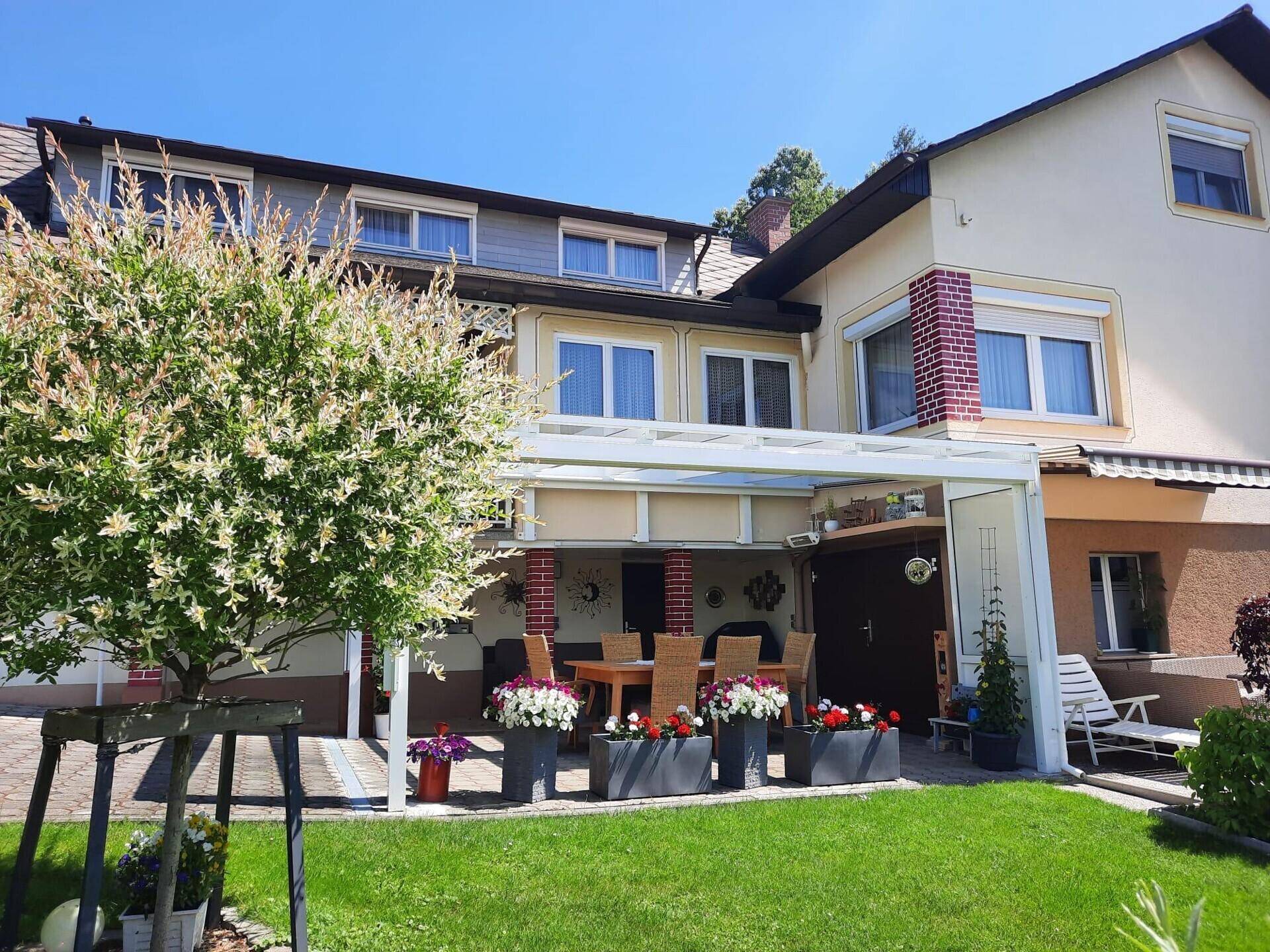 Gartenansicht des Hauses mit überdachter Terrasse, Esstisch und Blumenkübeln.